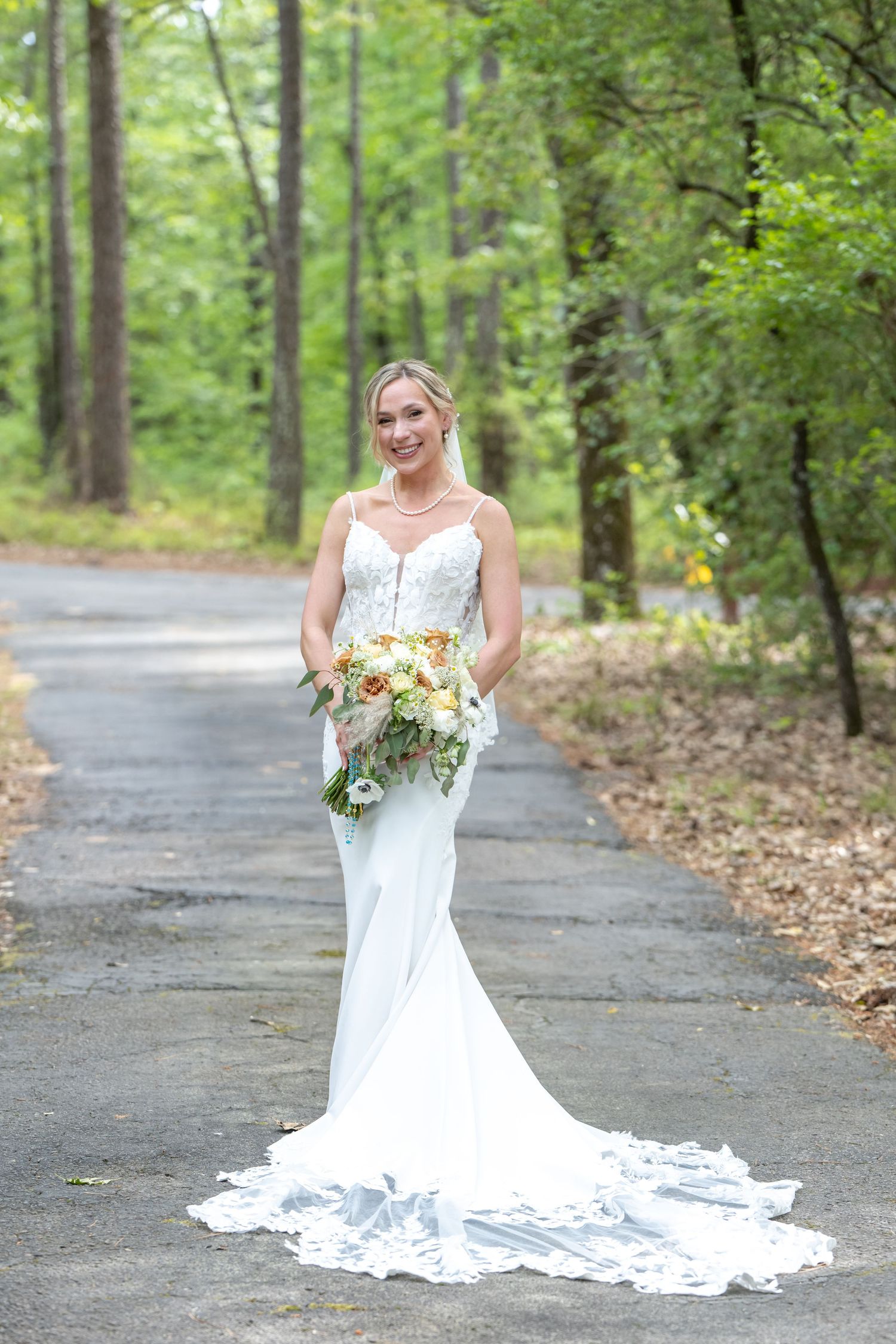 Bride in white mermaid gown with lace train holds bouquet on tree-lined path.