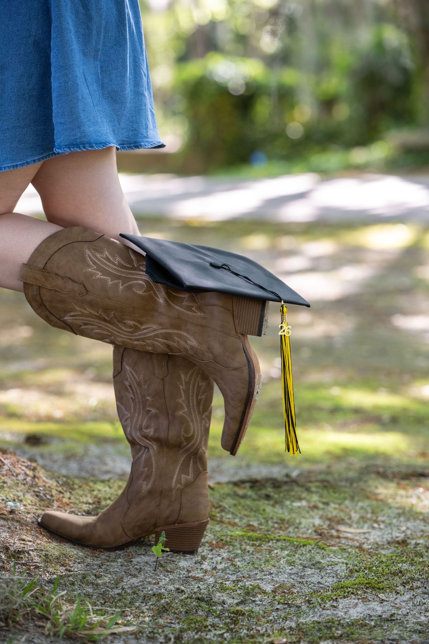 Person wearing cowboy boots and denim skirt holding a black graduation cap with gold tassel outdoors.