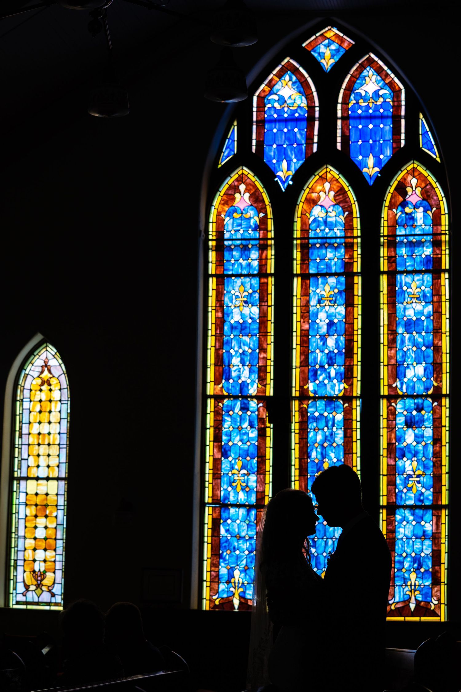 Silhouette bride and groom against colorful stained glass church windows.