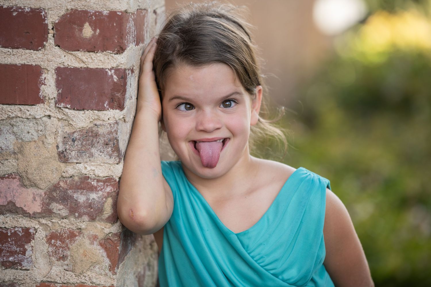 Young girl in teal dress sticking out tongue, leaning against rustic brick wall outdoors.