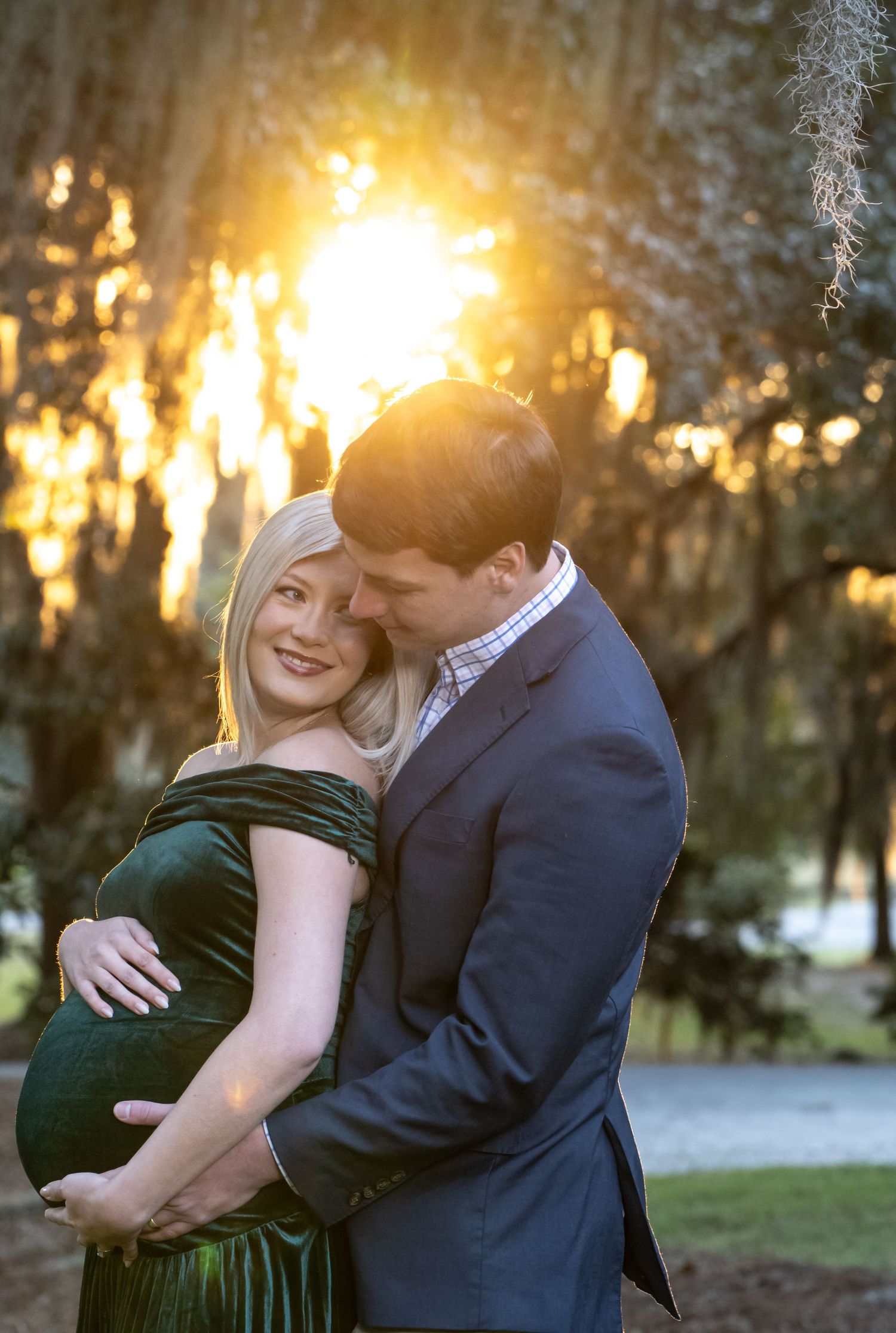 Couple embracing at golden hour sunset, pregnant woman in green velvet dress with partner in navy suit among moss-draped trees.