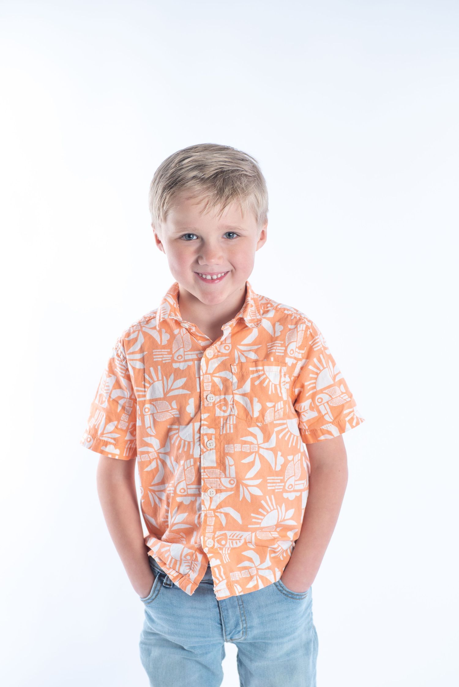 Young child wearing an orange tropical print button-down shirt and jeans stands with hands in pockets against white background.