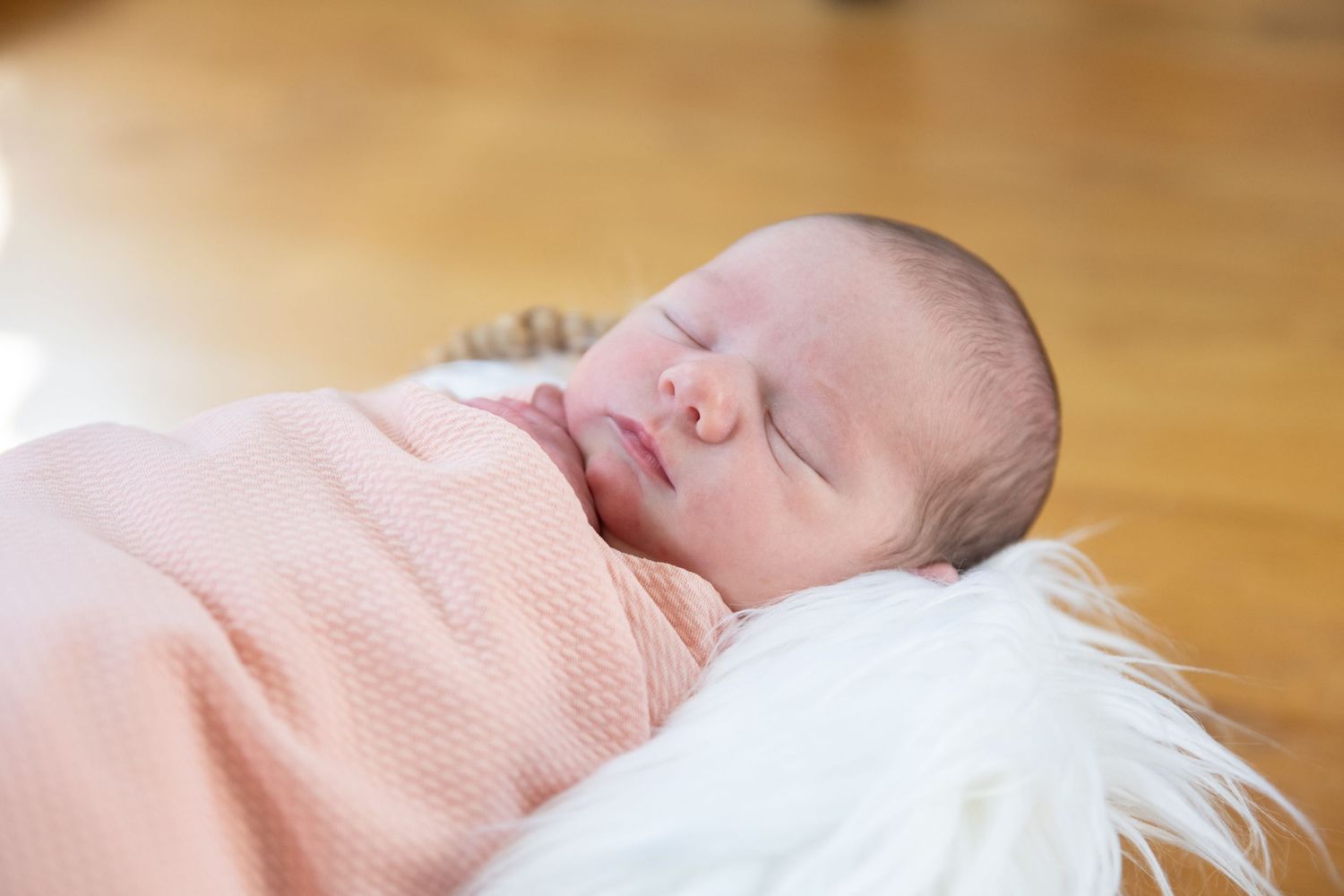 A peaceful sleeping newborn swaddled in a soft peach blanket rests on white fluffy fur.