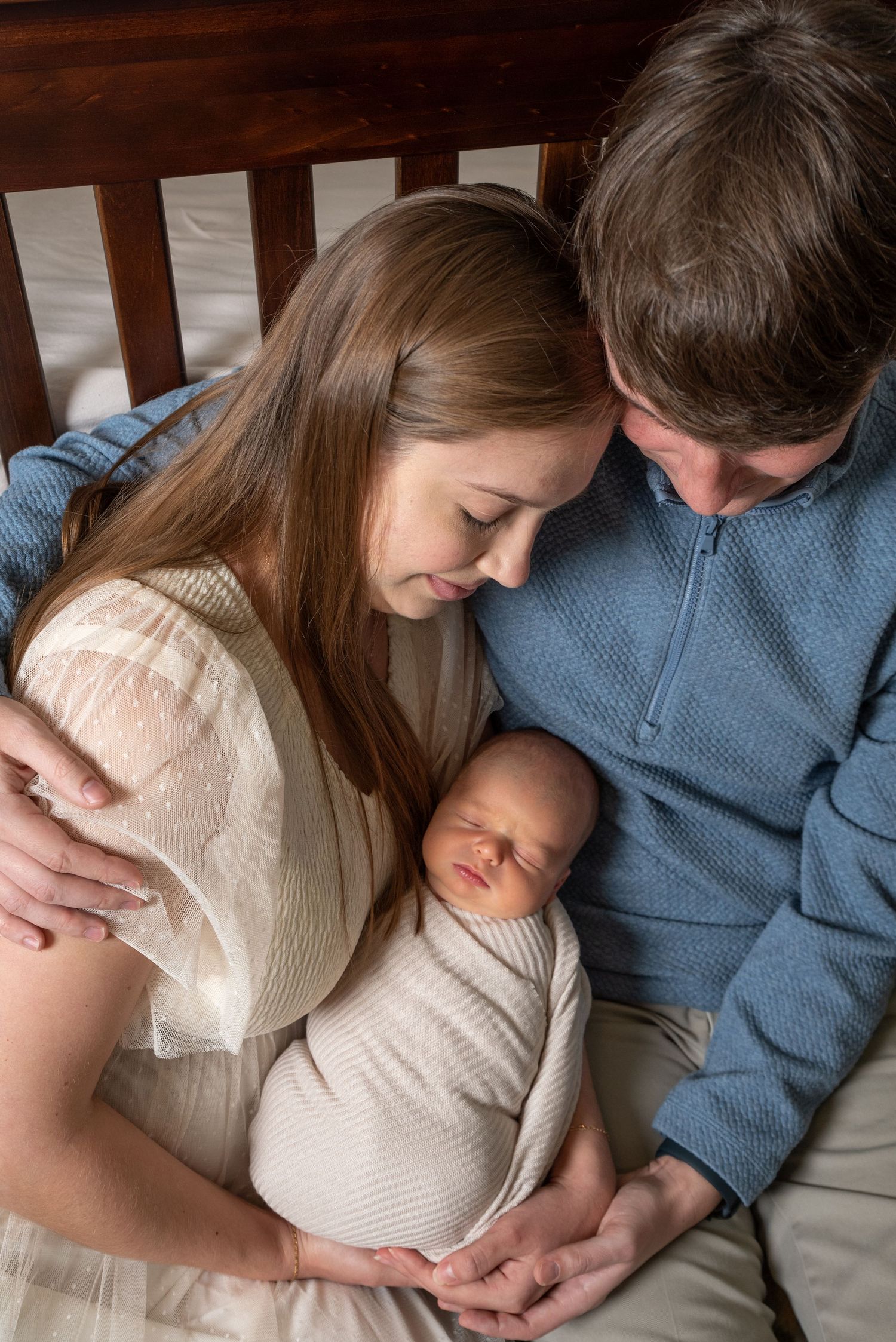 New parents tenderly gaze at their swaddled newborn baby while sitting together on a bed.