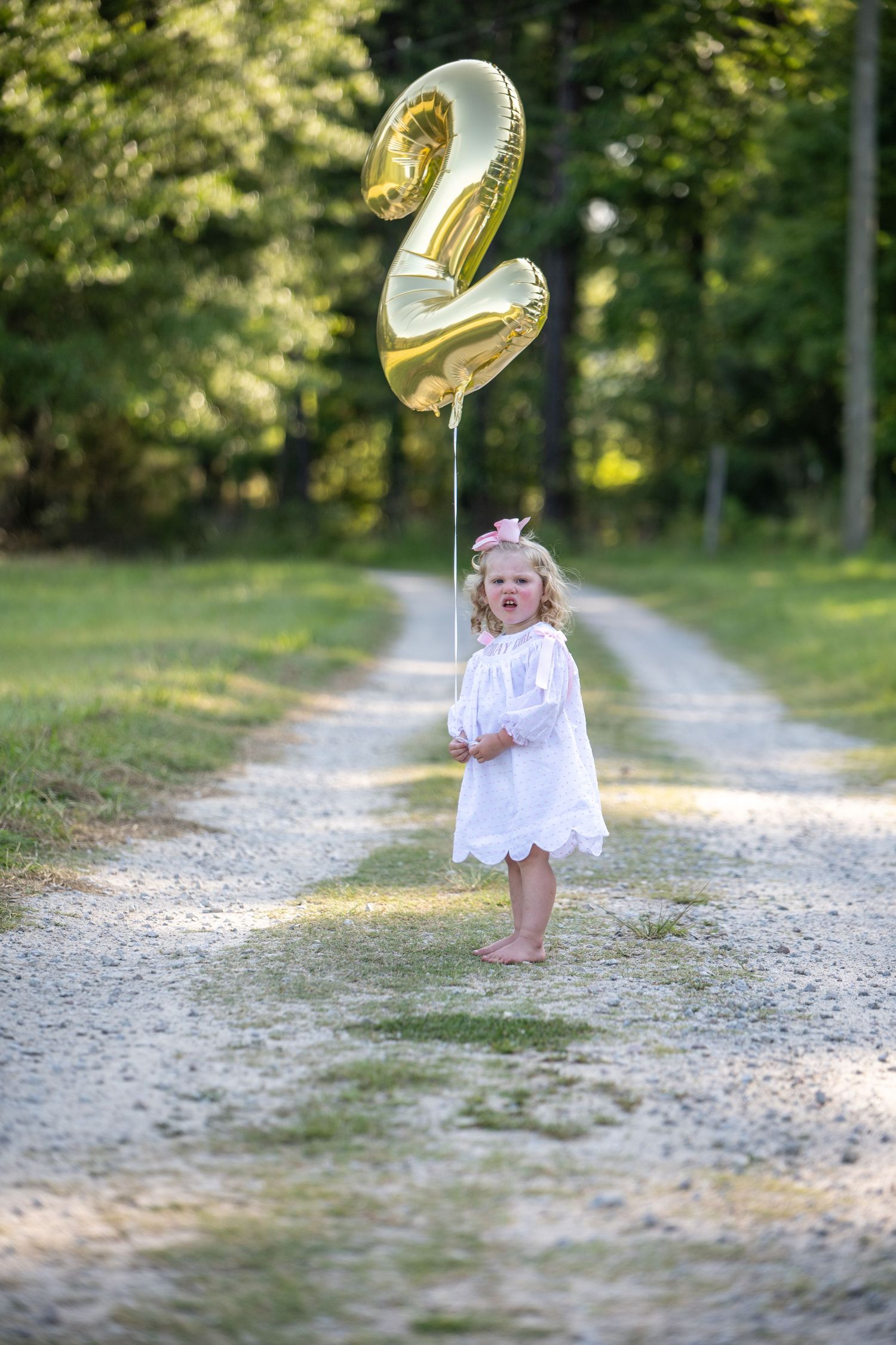 Toddler girl in white dress holding gold number 2 balloon on gravel path surrounded by green trees.