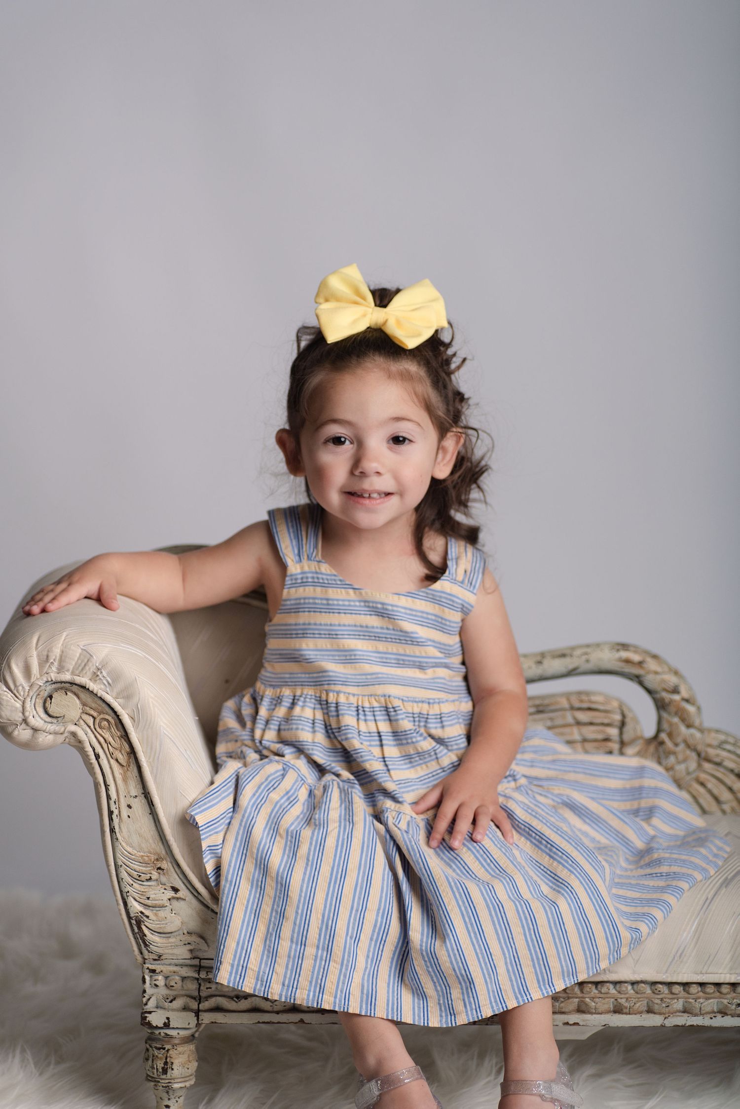 Young girl in striped dress with yellow bow sits on vintage chaise lounge, smiling for portrait photo.
