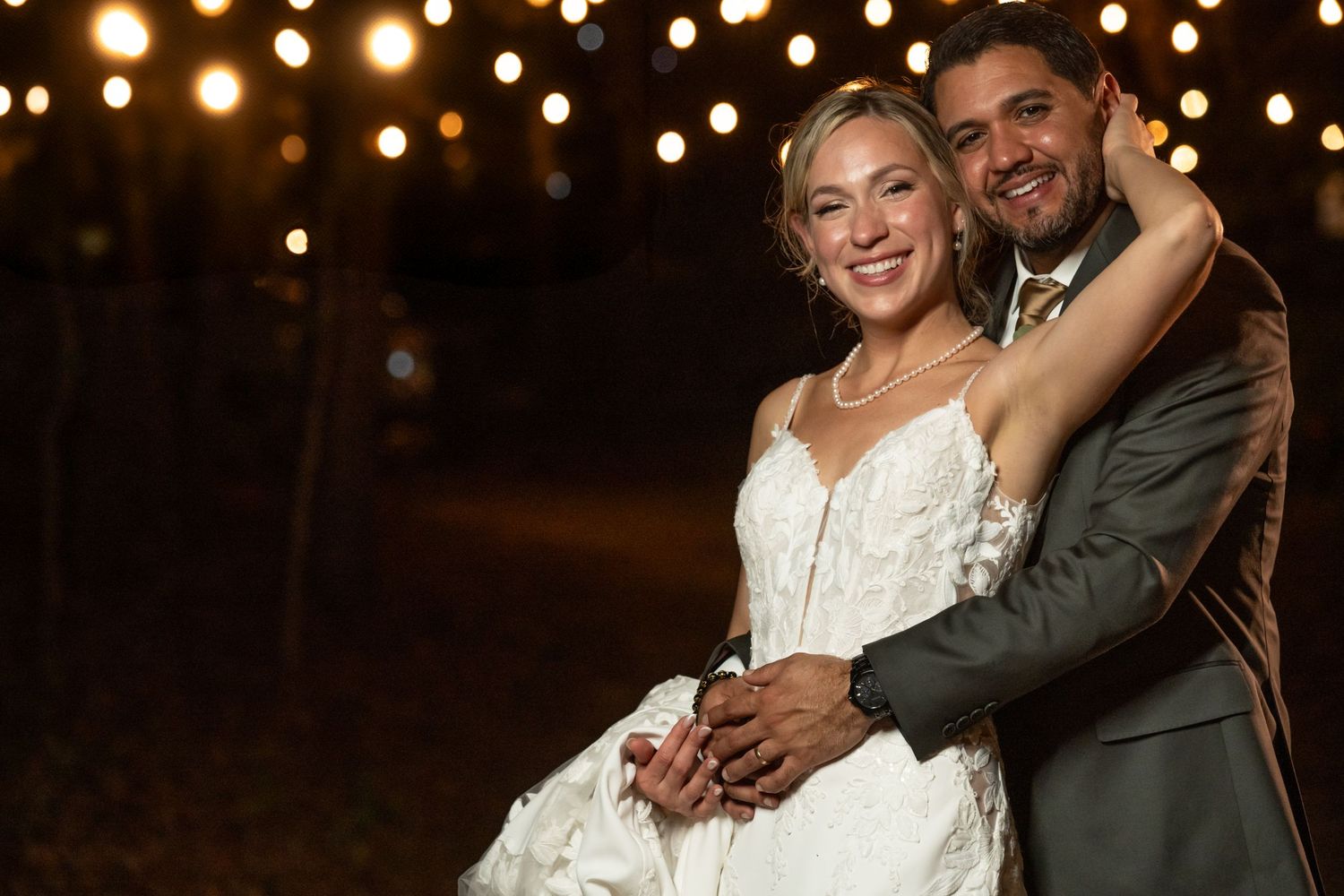 Bride and groom embrace under string lights at night, smiling joyfully in wedding attire.
