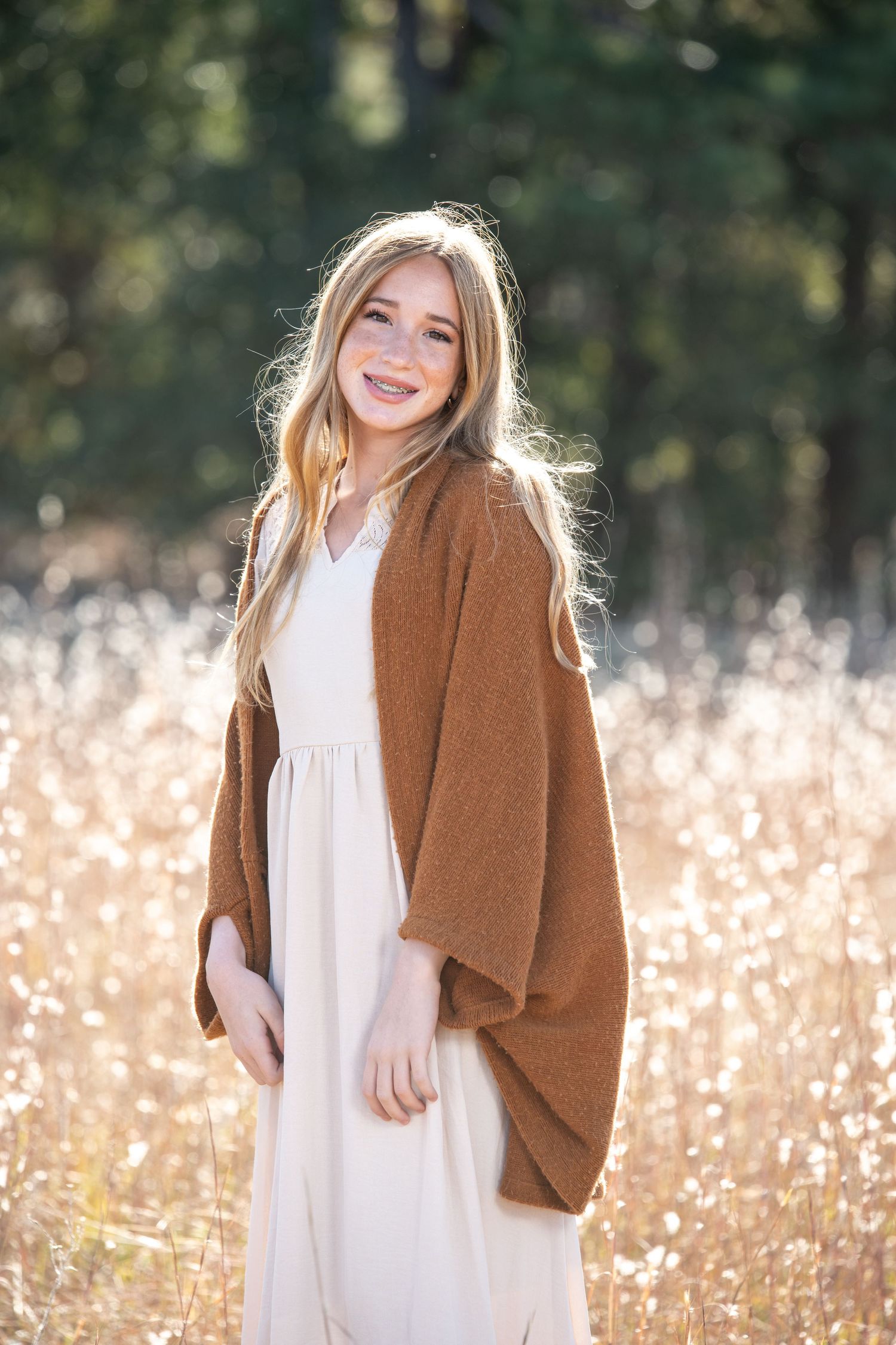 Young blonde woman smiling in a white dress and brown cardigan, standing in a sunlit golden field with trees behind her.