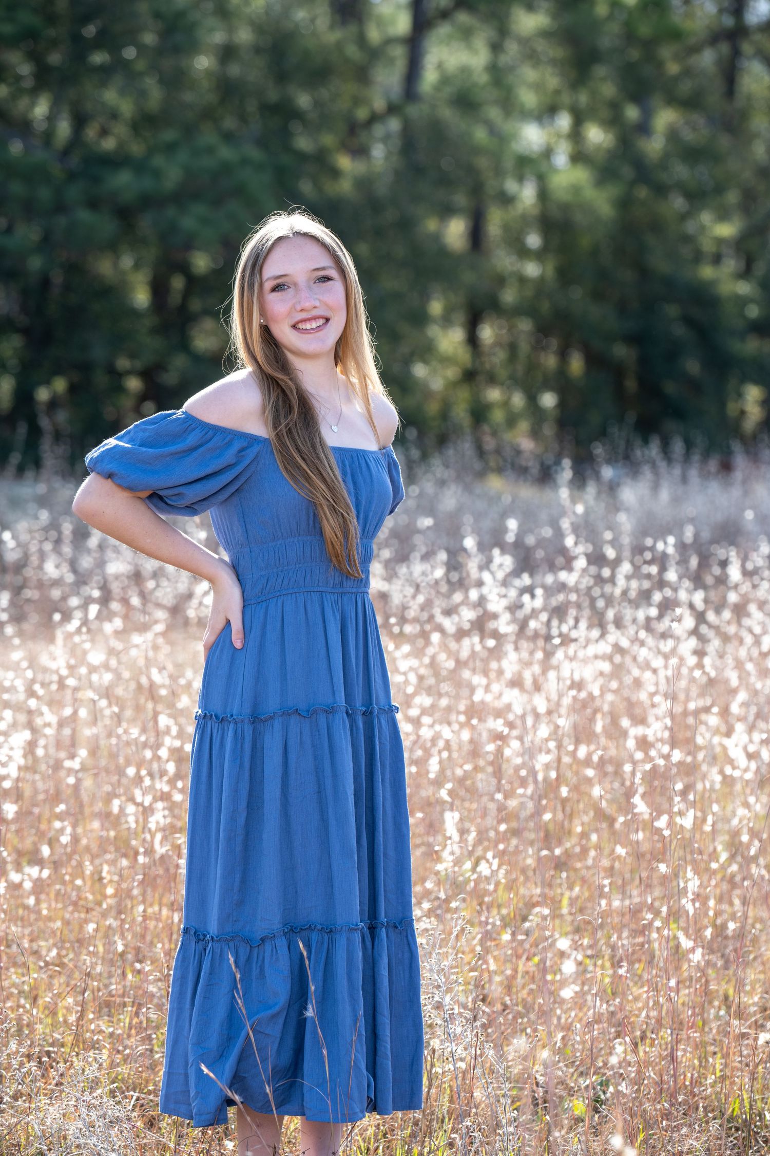 Young woman in blue off-shoulder tiered maxi dress smiling in a sunlit field with trees in background.