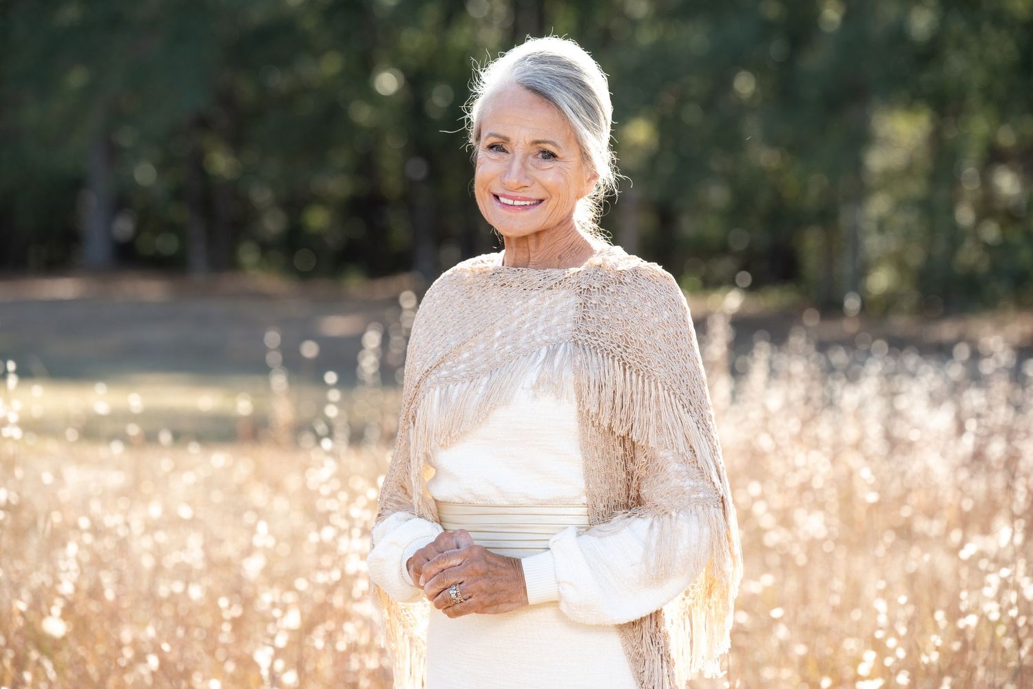 Elegant older woman with silver hair wearing a white dress and beaded cape, standing in a golden field.