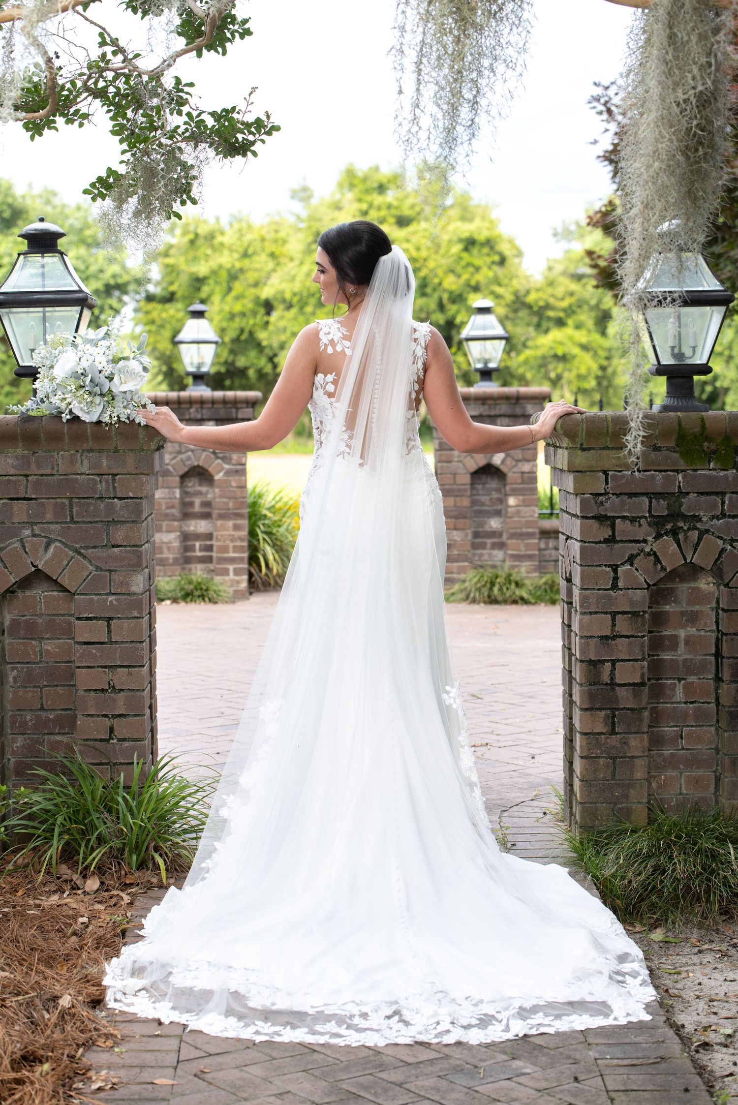Bride in lace wedding gown with cathedral veil holding bouquet, standing between brick pillars in lush garden setting.