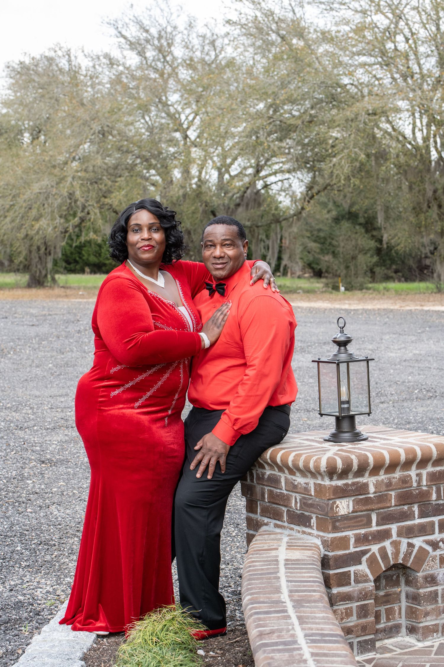 A couple wearing coordinating red outfits poses together by a brick wall and lantern outdoors on a winter day.