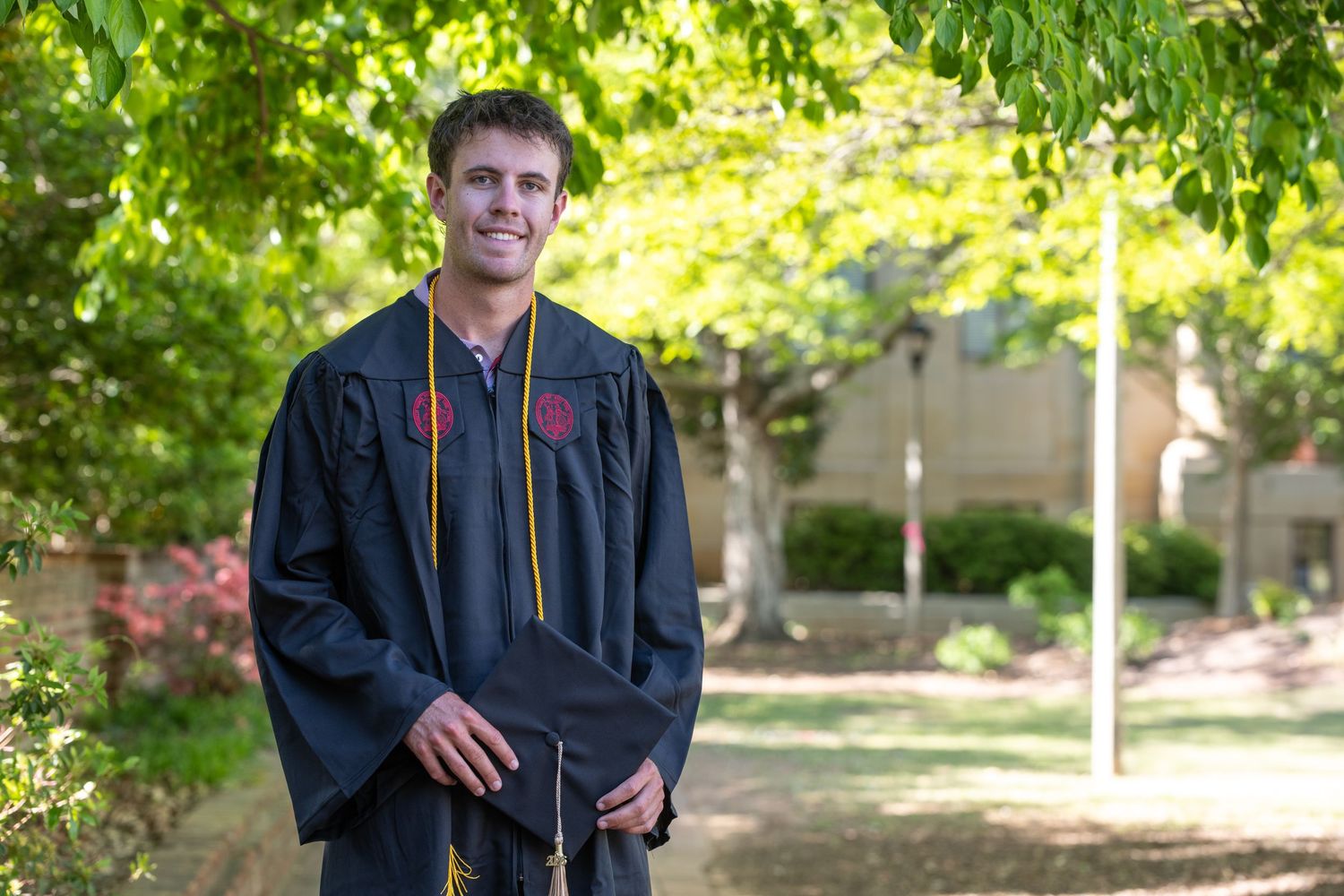 Young man in graduation gown and stole smiling outdoors on a sunny campus surrounded by green trees.