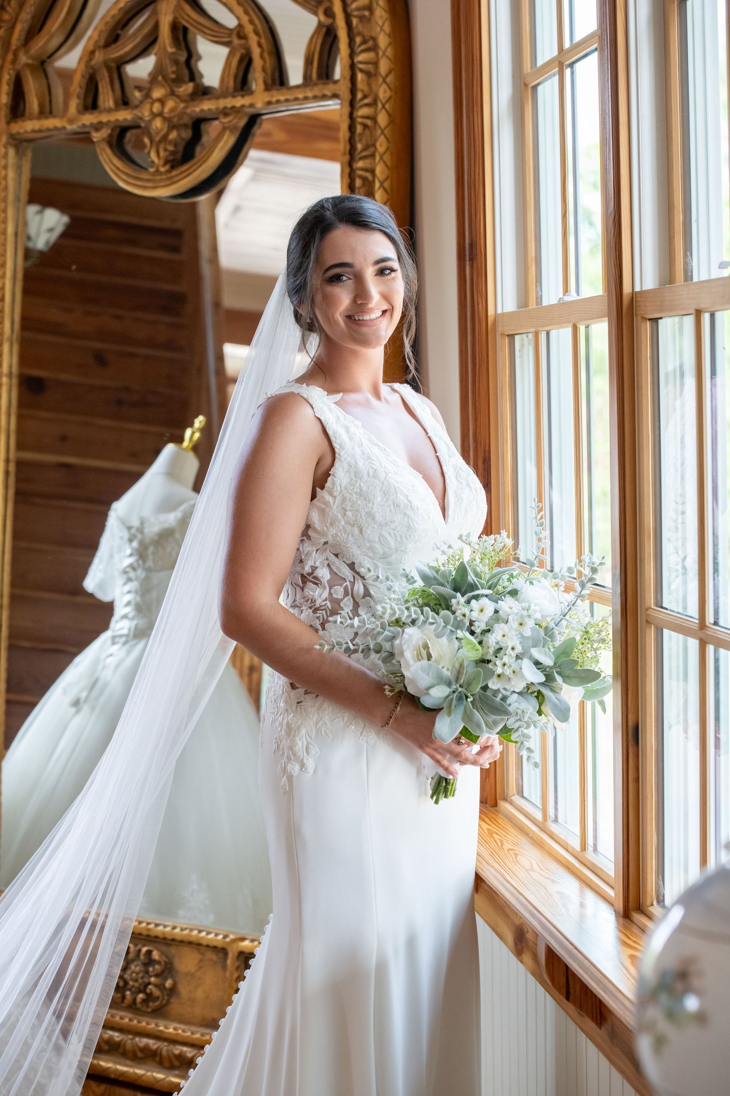 Smiling bride in lace wedding gown holding white floral bouquet, standing by sunlit window with veil flowing.
