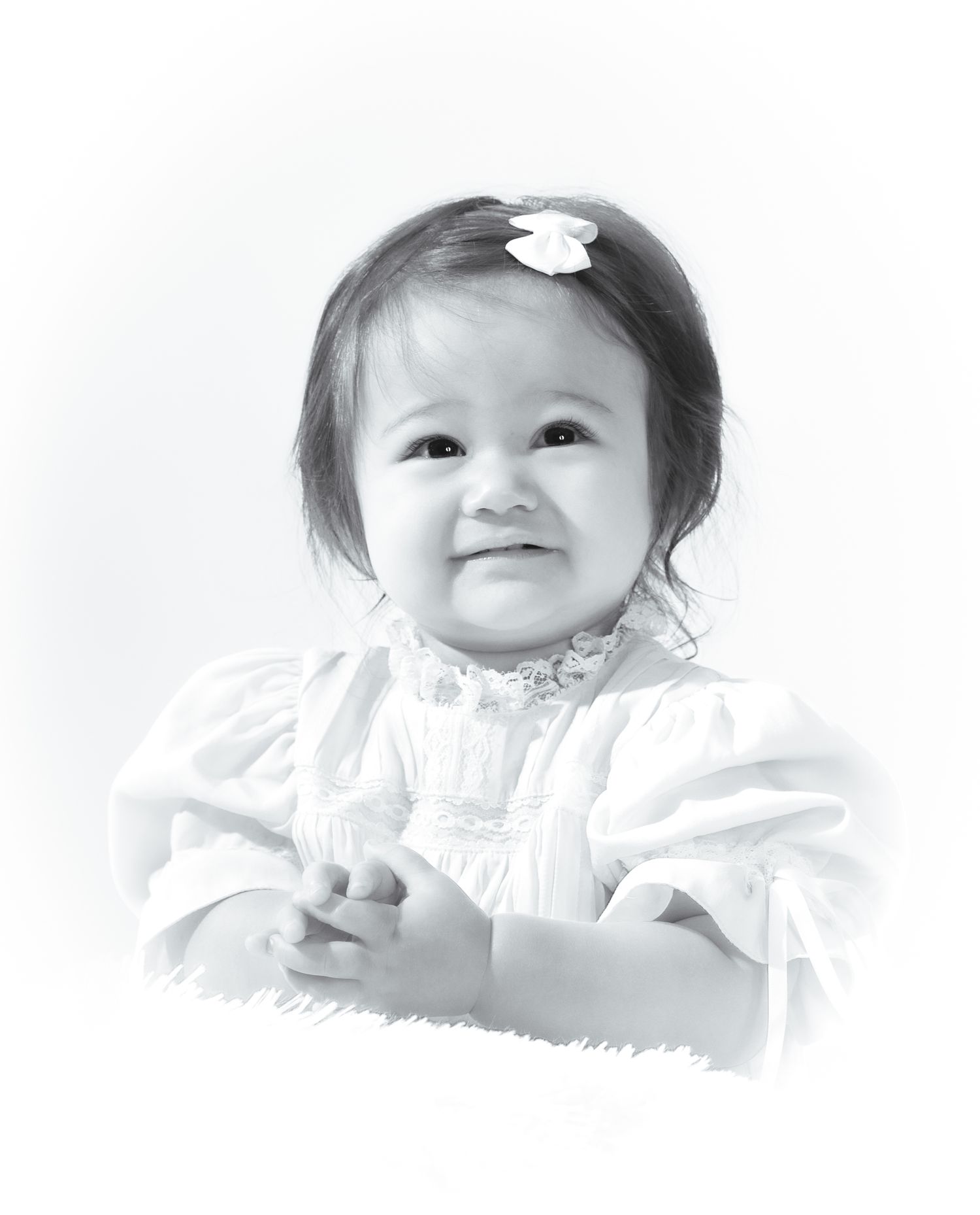 Black and white portrait of a smiling baby girl with a white bow in her dark hair, wearing a white dress.
