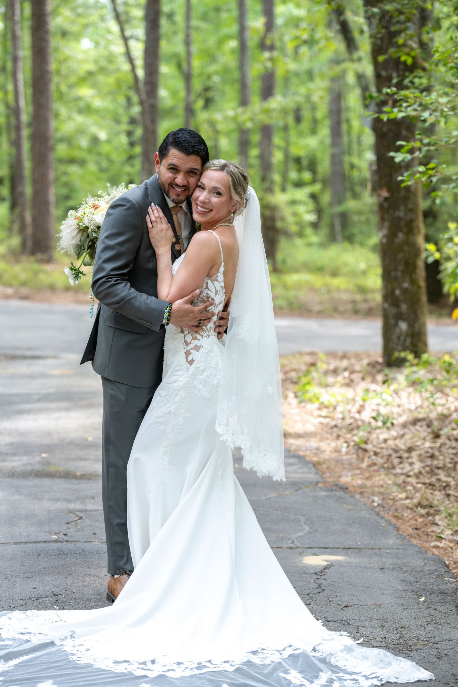 Smiling bride and groom pose on a tree-lined path, bride in lace gown with veil, groom in gray suit holding bouquet.