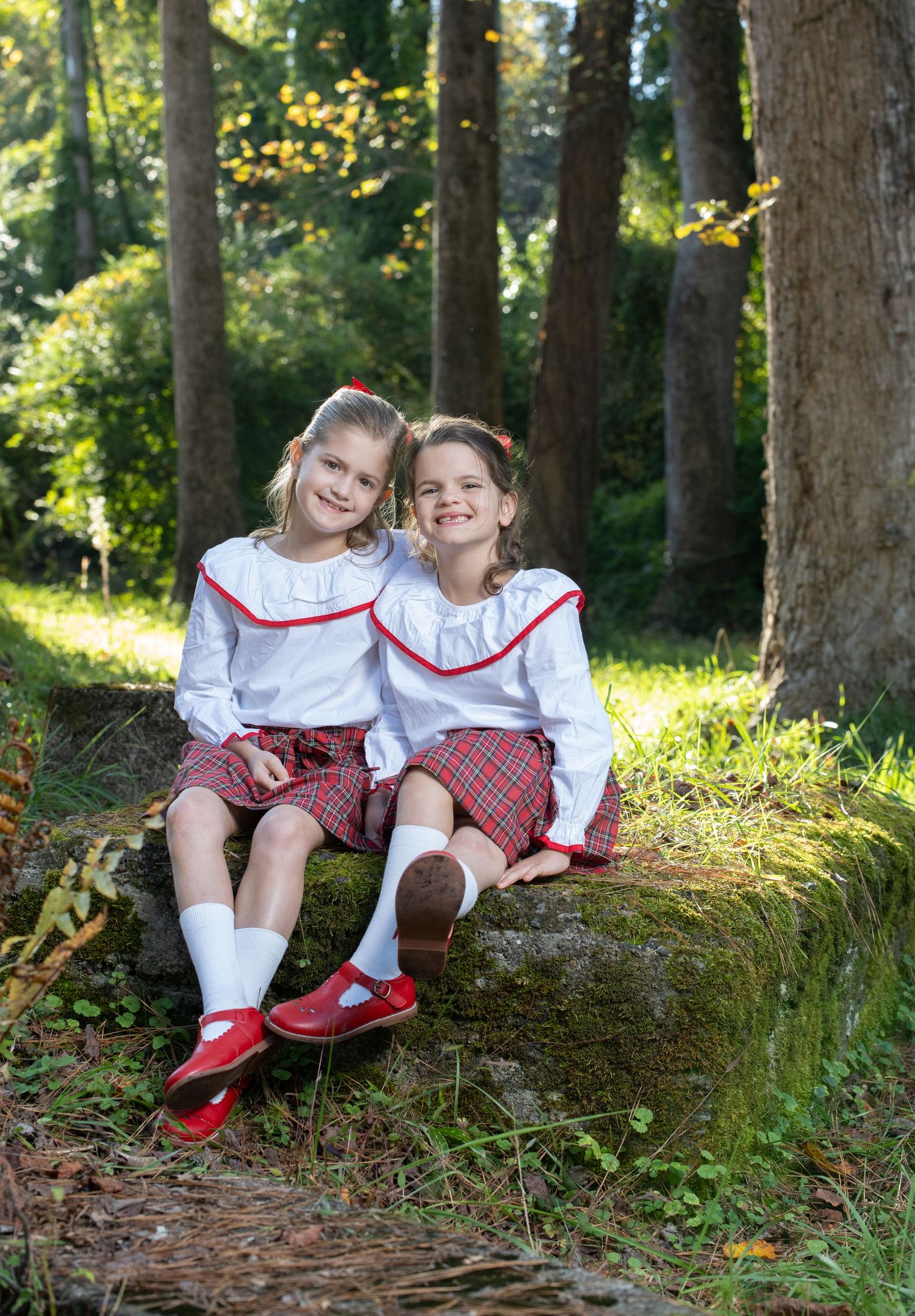 Two girls in matching red plaid shorts and white blouses with red trim sit together on a rock in a sunny forest.