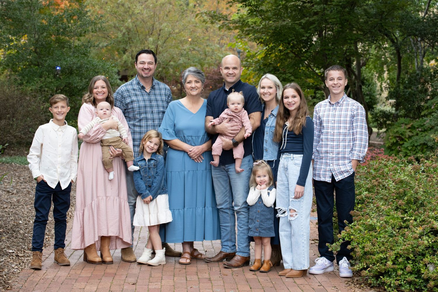 Large family group portrait smiling outdoors on brick path surrounded by autumn trees.