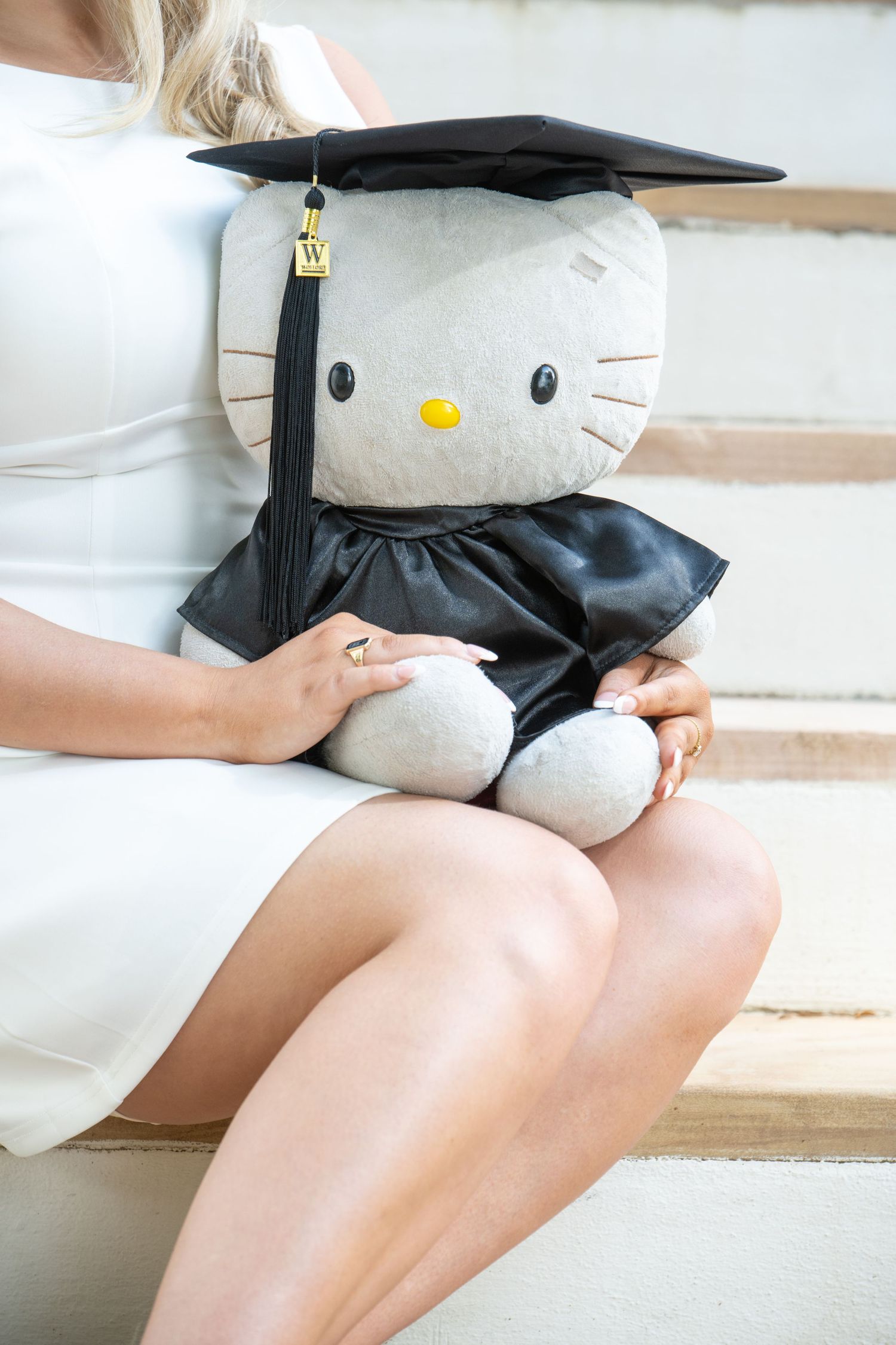Hello Kitty plush toy wearing graduation cap and gown held by woman in white dress on steps.