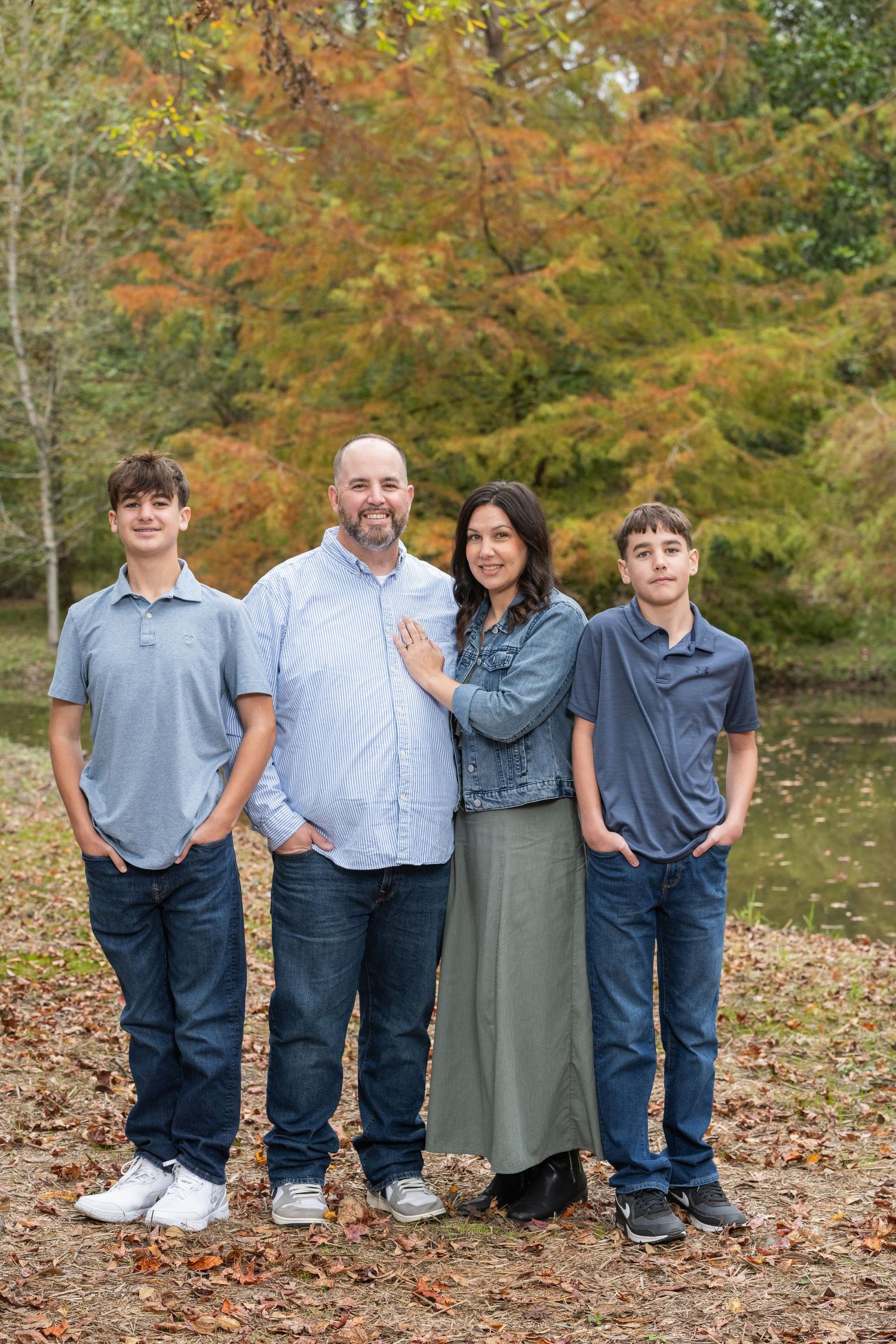 Family of four smiling outdoors in autumn, standing near a pond surrounded by colorful fall foliage.