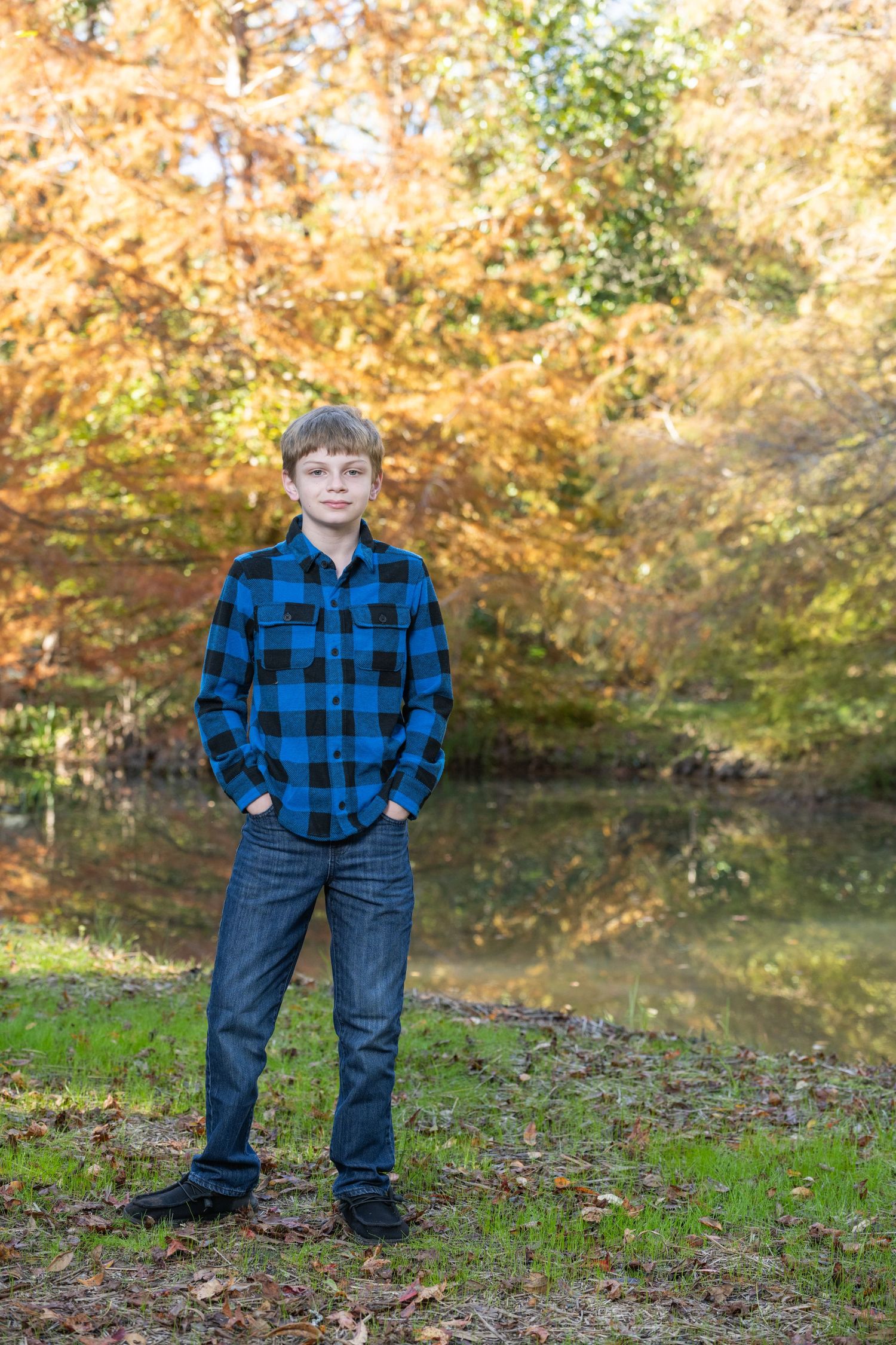 A young boy in a blue plaid flannel shirt and jeans stands outdoors near a pond surrounded by colorful autumn foliage.