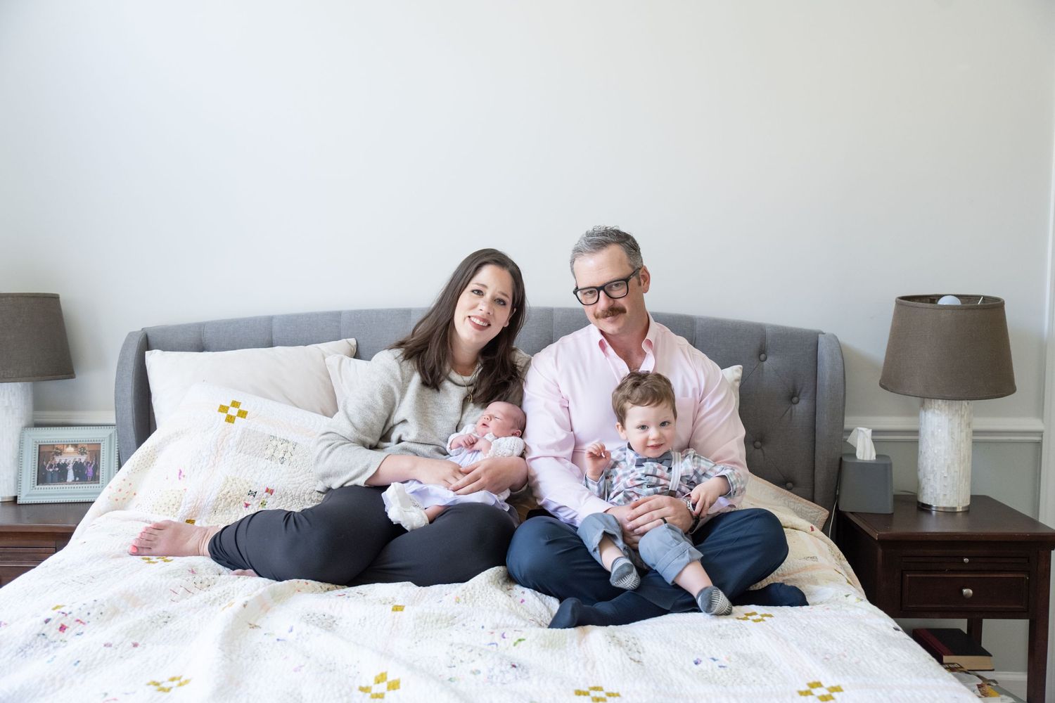 A family sits together on a gray bed with matching lamps and nightstands in a bright, modern bedroom.