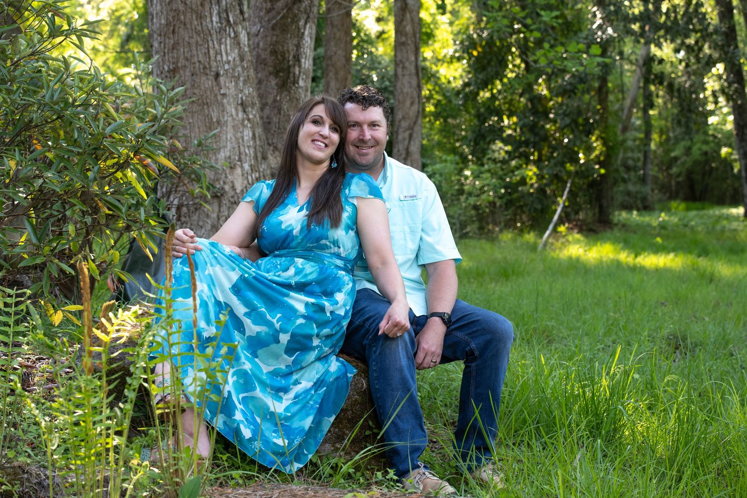 Couple smiling outdoors in lush green forest, woman in blue floral dress, man in light blue shirt and jeans.