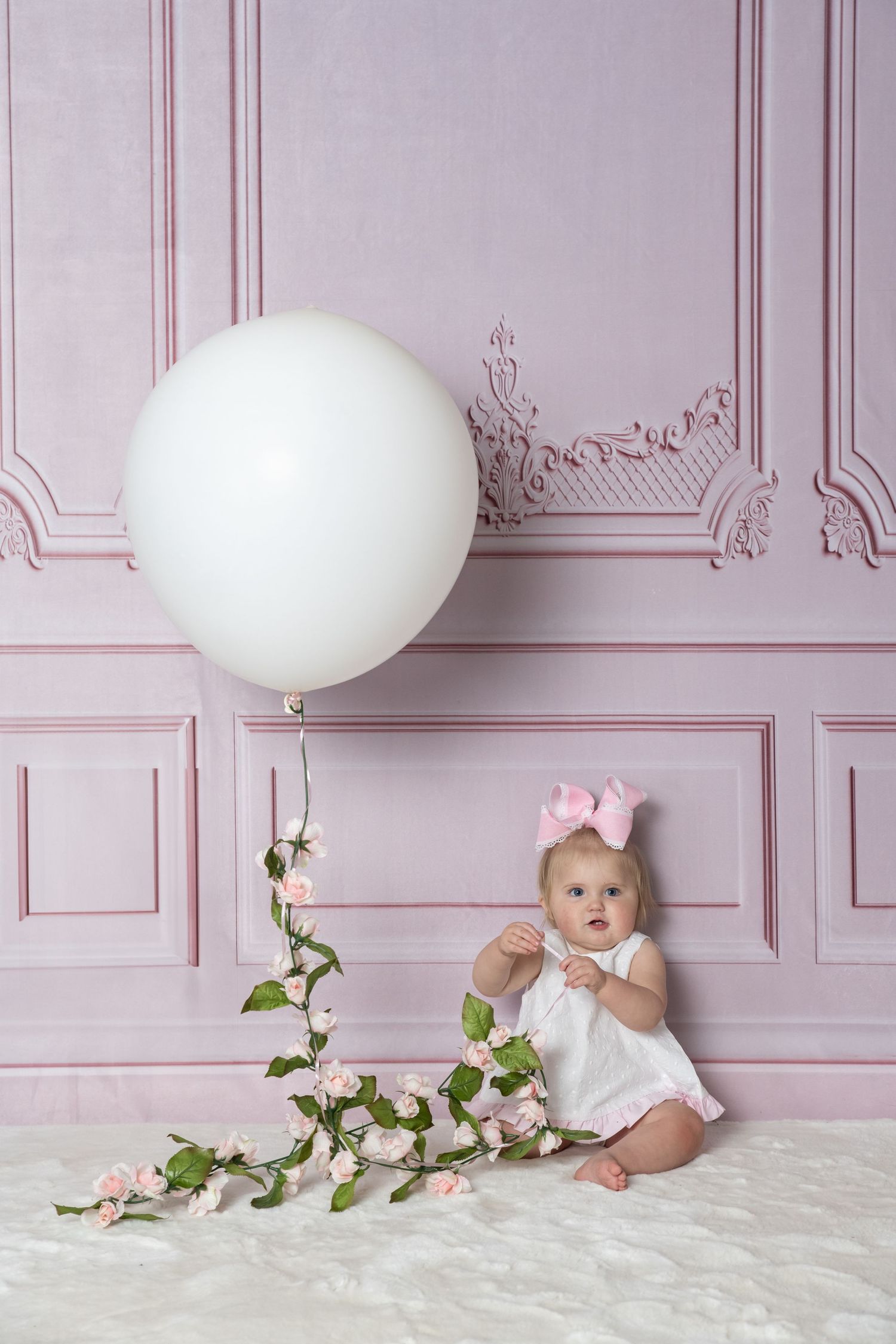 Baby in white dress sits against pink wall with decorative white balloon and floral garland.