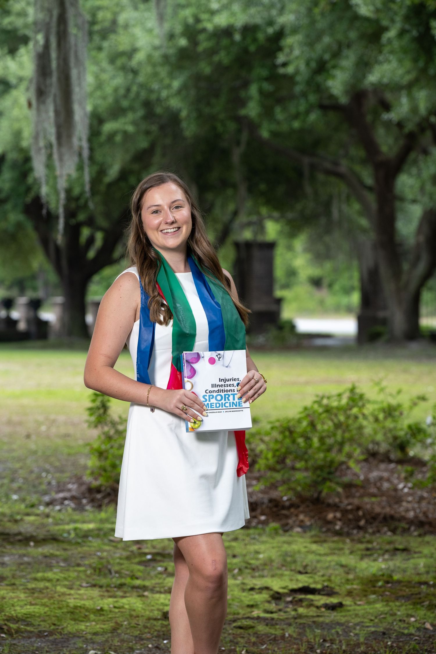 Smiling young woman in white dress with colorful sash holding a sports booklet outdoors among green trees.