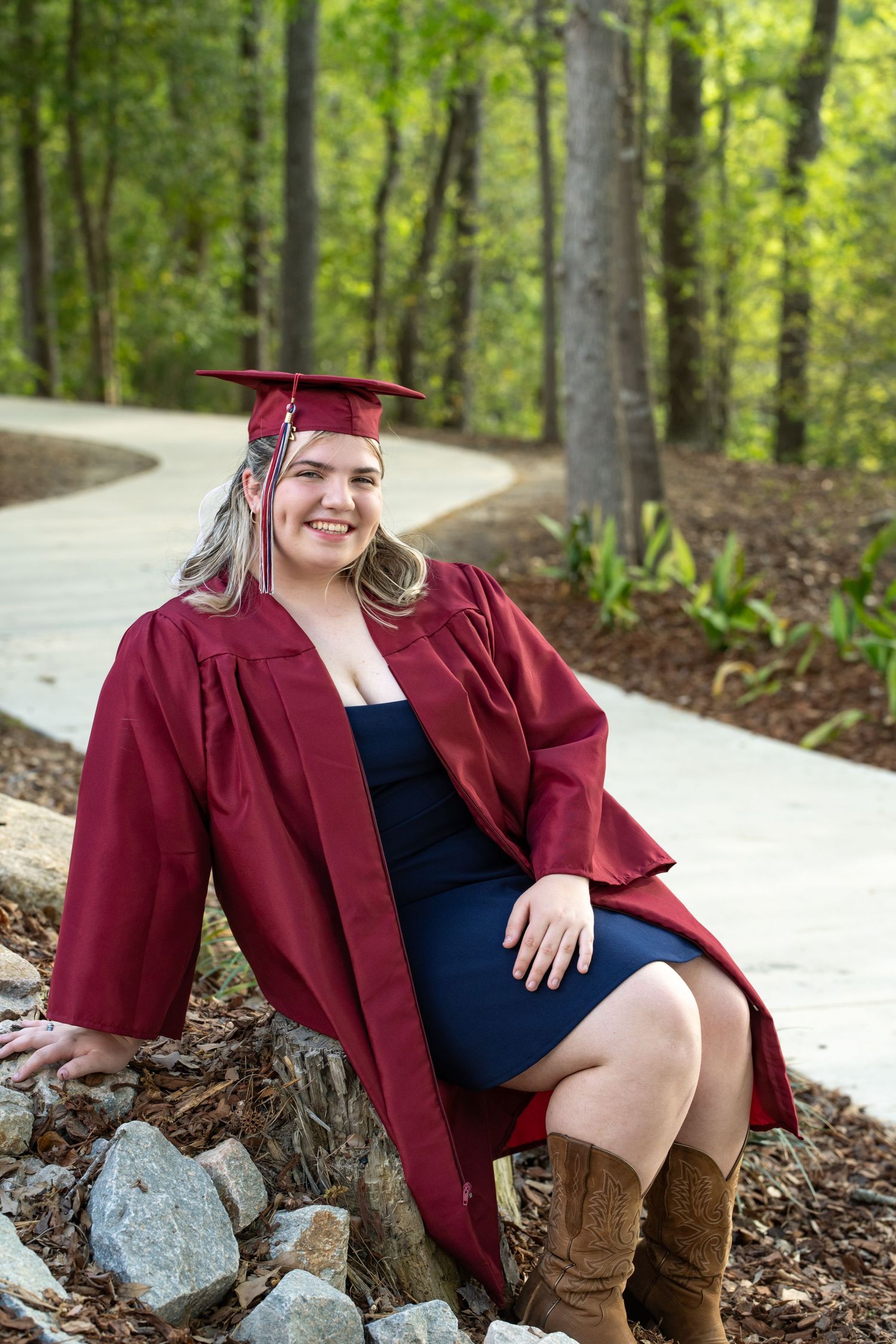 Smiling young woman in maroon graduation cap and gown sitting on rocks along a wooded pathway.
