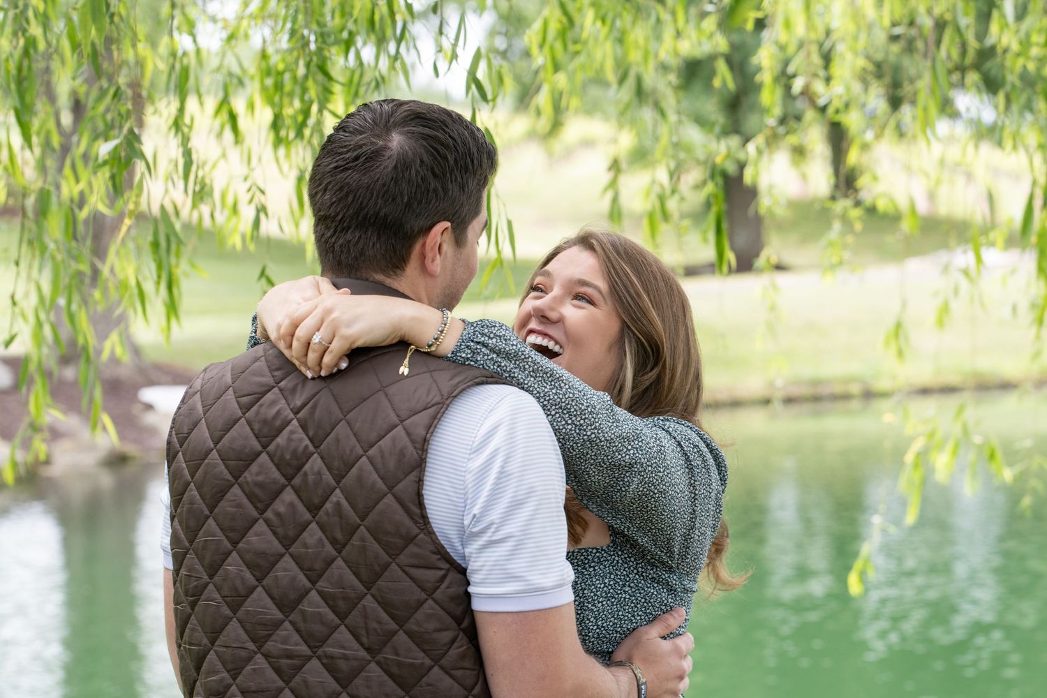 Happy couple embracing and laughing together near a serene green pond under a weeping willow tree.