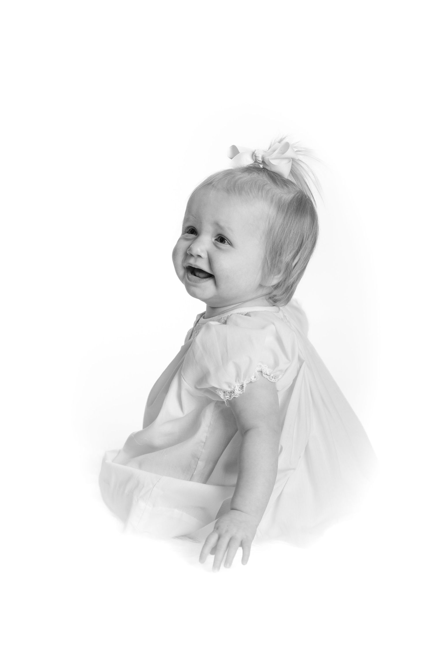 Black and white portrait of a smiling baby girl in a white dress with a bow in her hair.