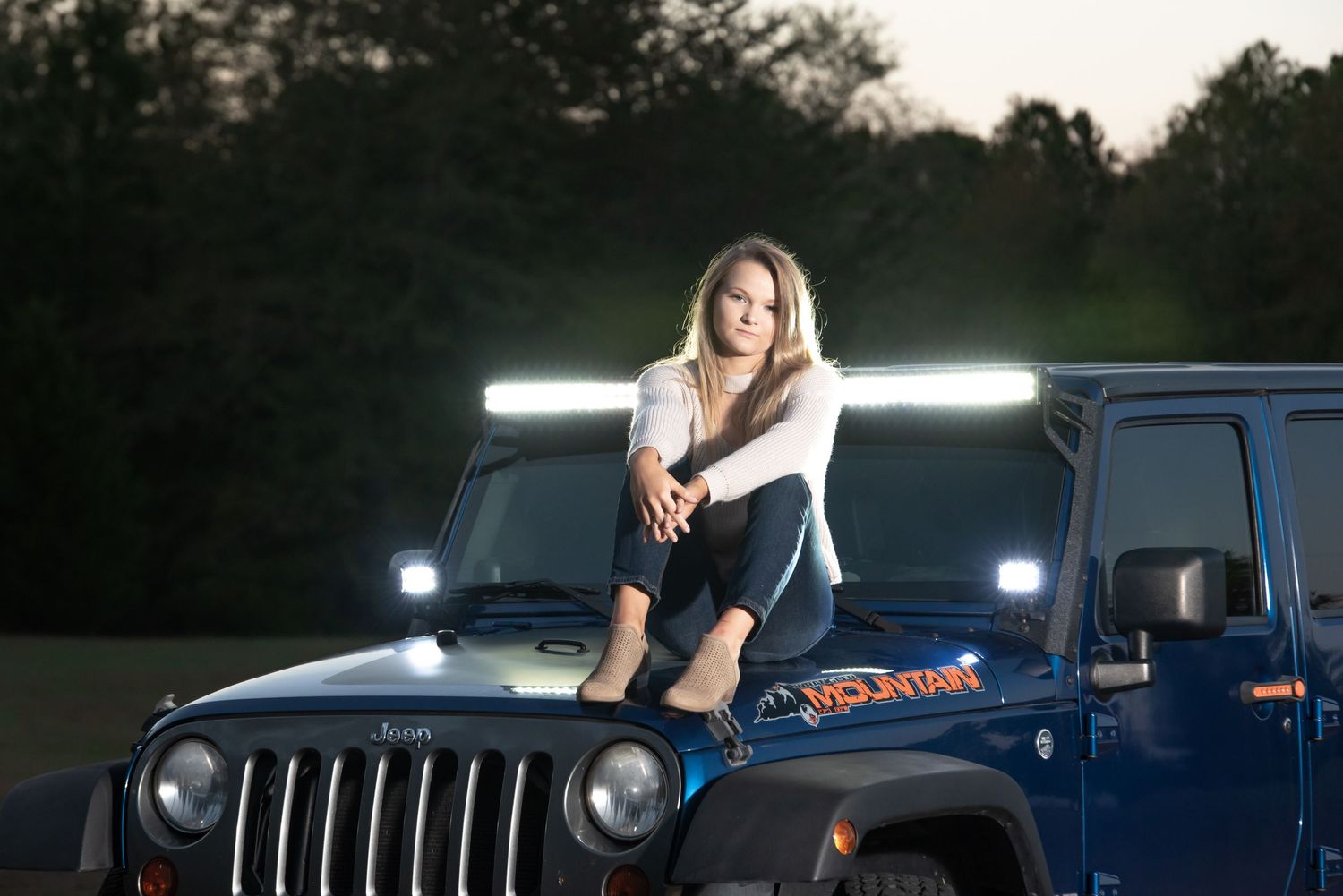 Woman poses on hood of blue Jeep with illuminated light bars at dusk.