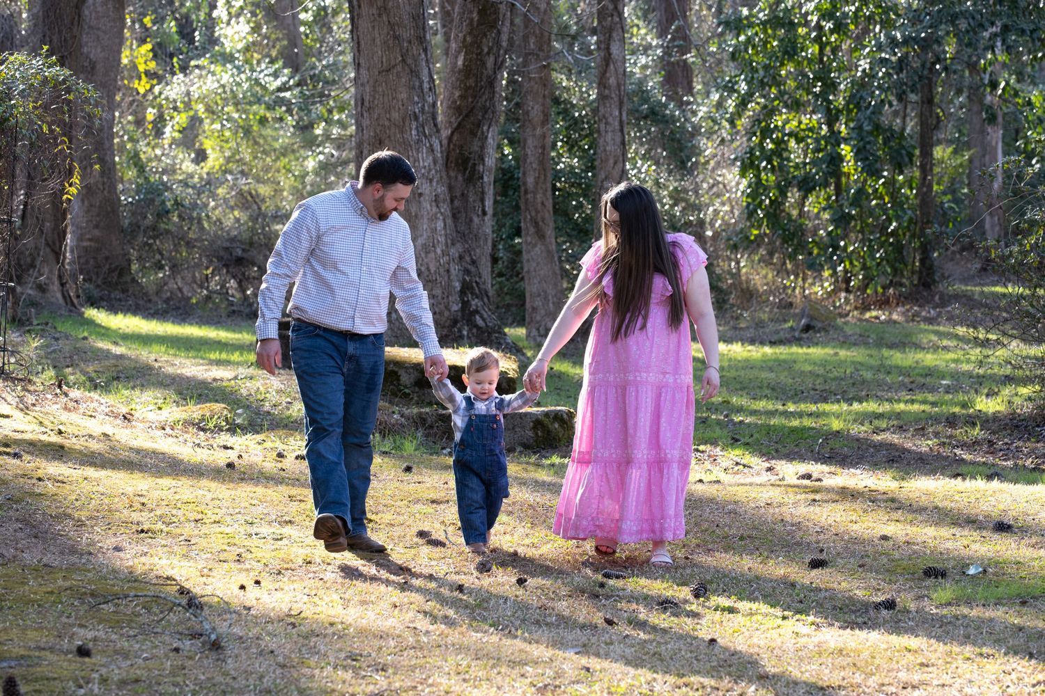 Family walking together on a sunlit wooded path while holding their child's hands in a scenic outdoor setting.