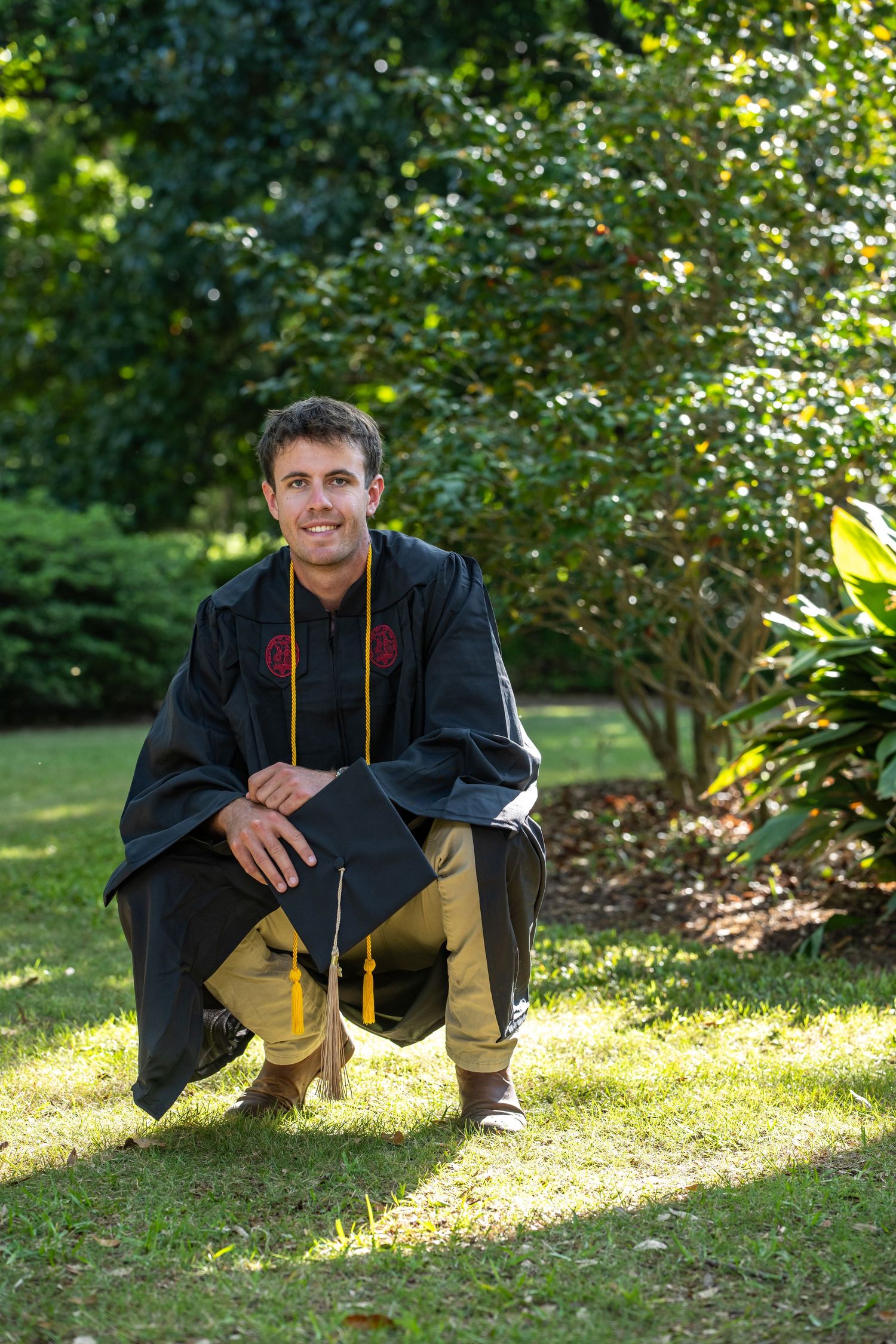 Young man in black graduation gown and gold tassel cap kneeling on grass in a sunny outdoor park setting.