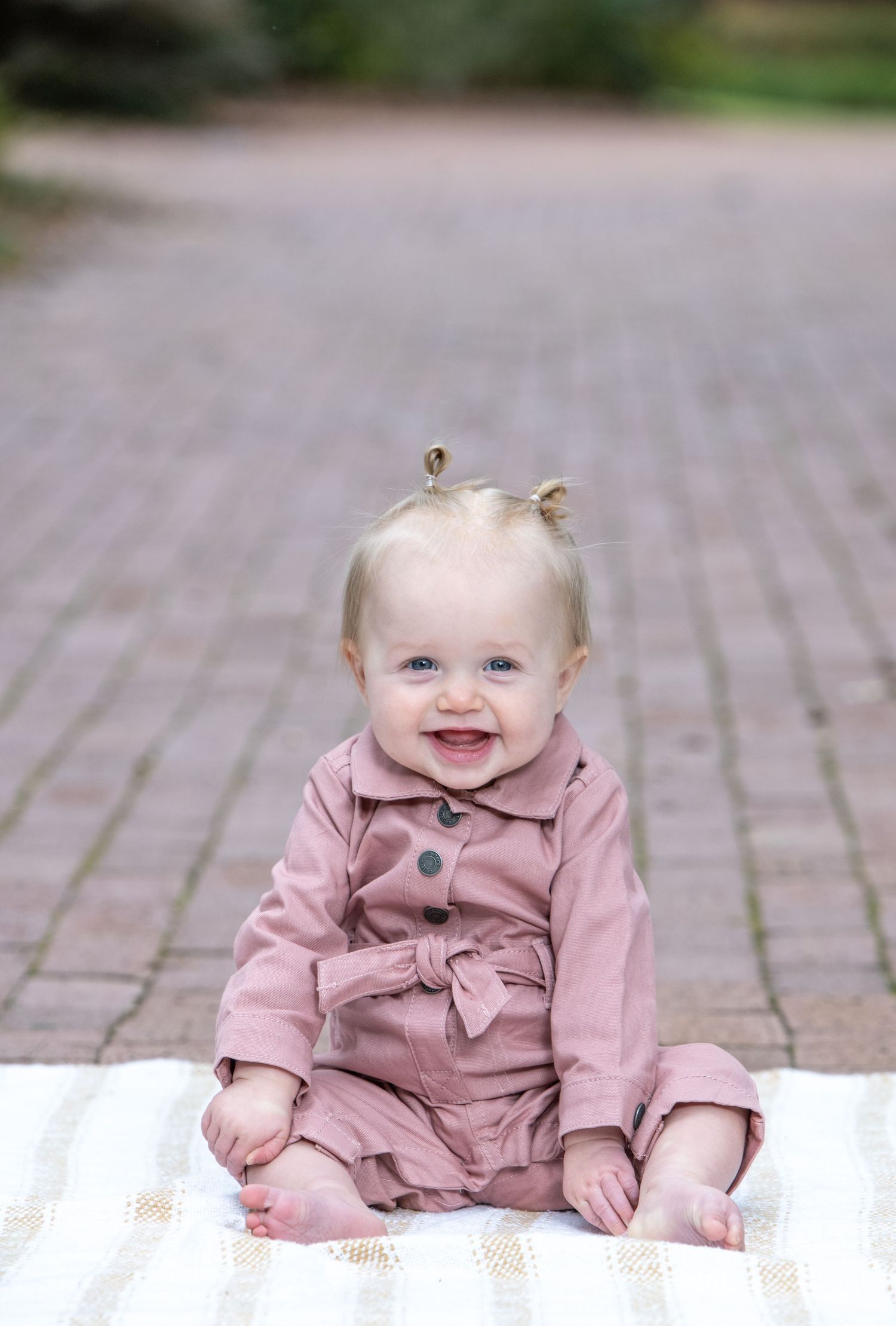 Smiling baby girl with blue eyes sitting on a blanket outdoors in a pink romper with tiny pigtails.