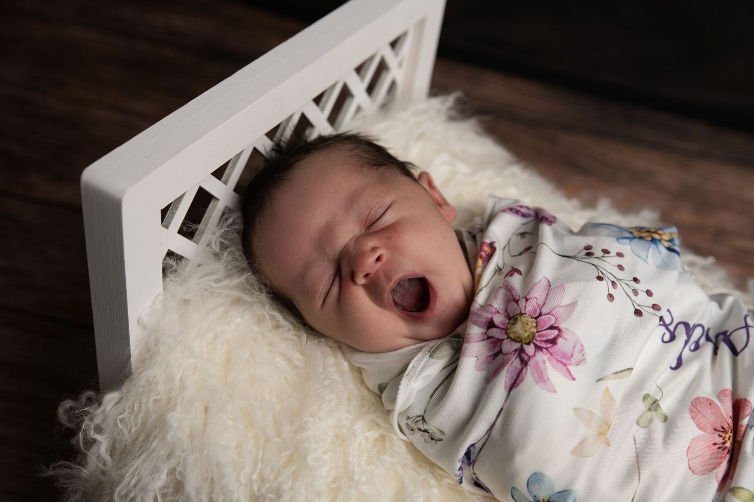 Newborn baby yawning while swaddled in floral blanket, sleeping in white mini crib with fluffy white fur lining.