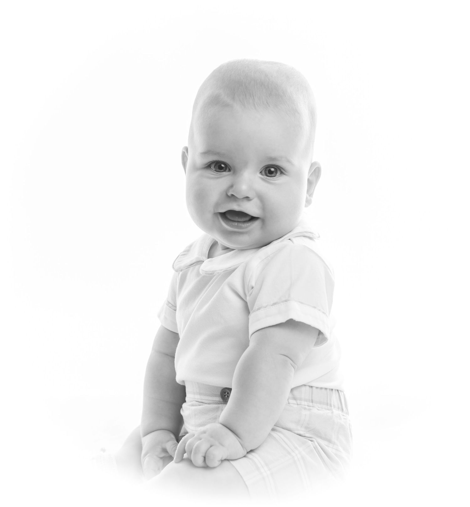 Black and white portrait of a smiling baby in white clothing, sitting upright against a white background.