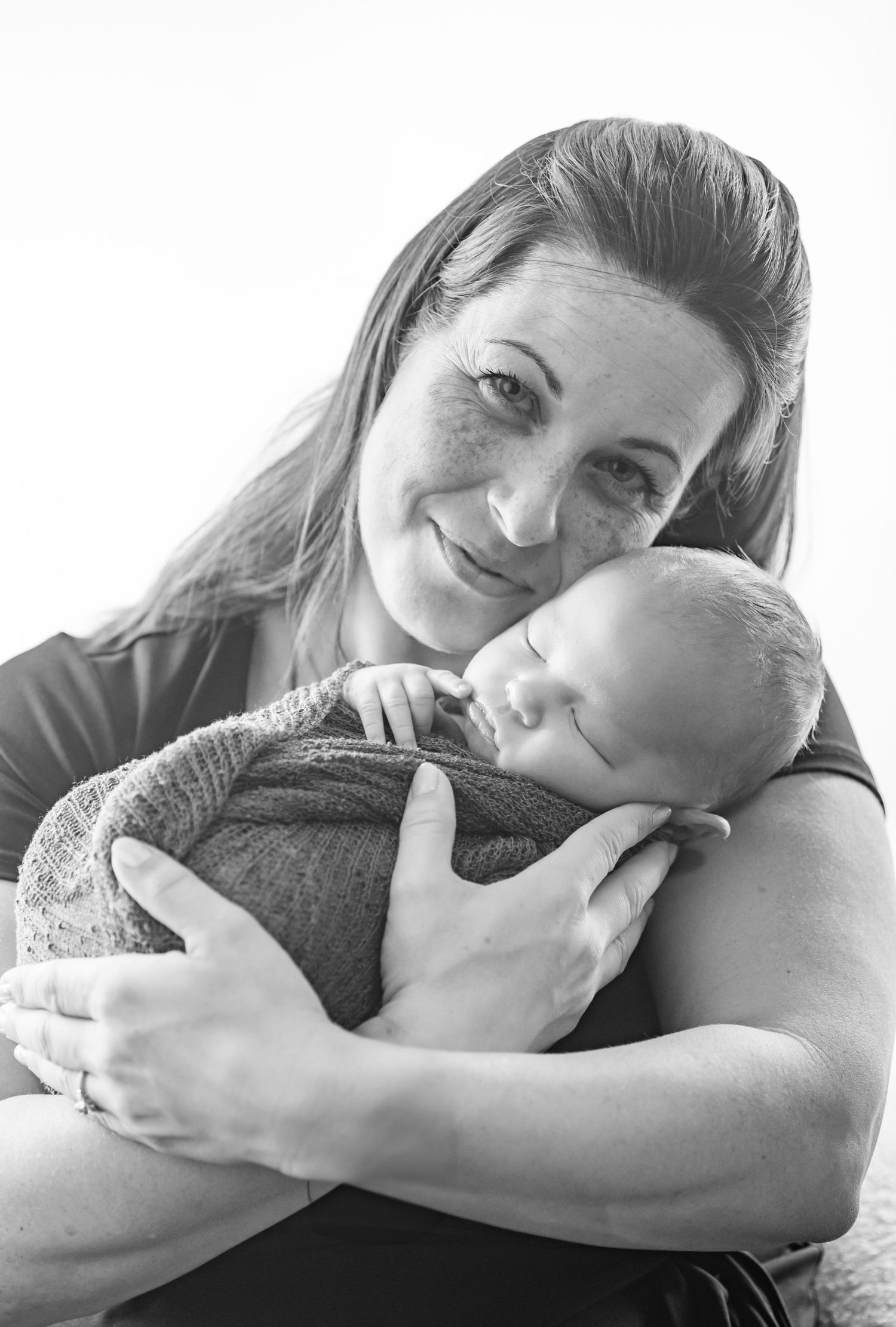 Black and white portrait of a smiling woman tenderly holding a sleeping newborn baby wrapped in a soft blanket.
