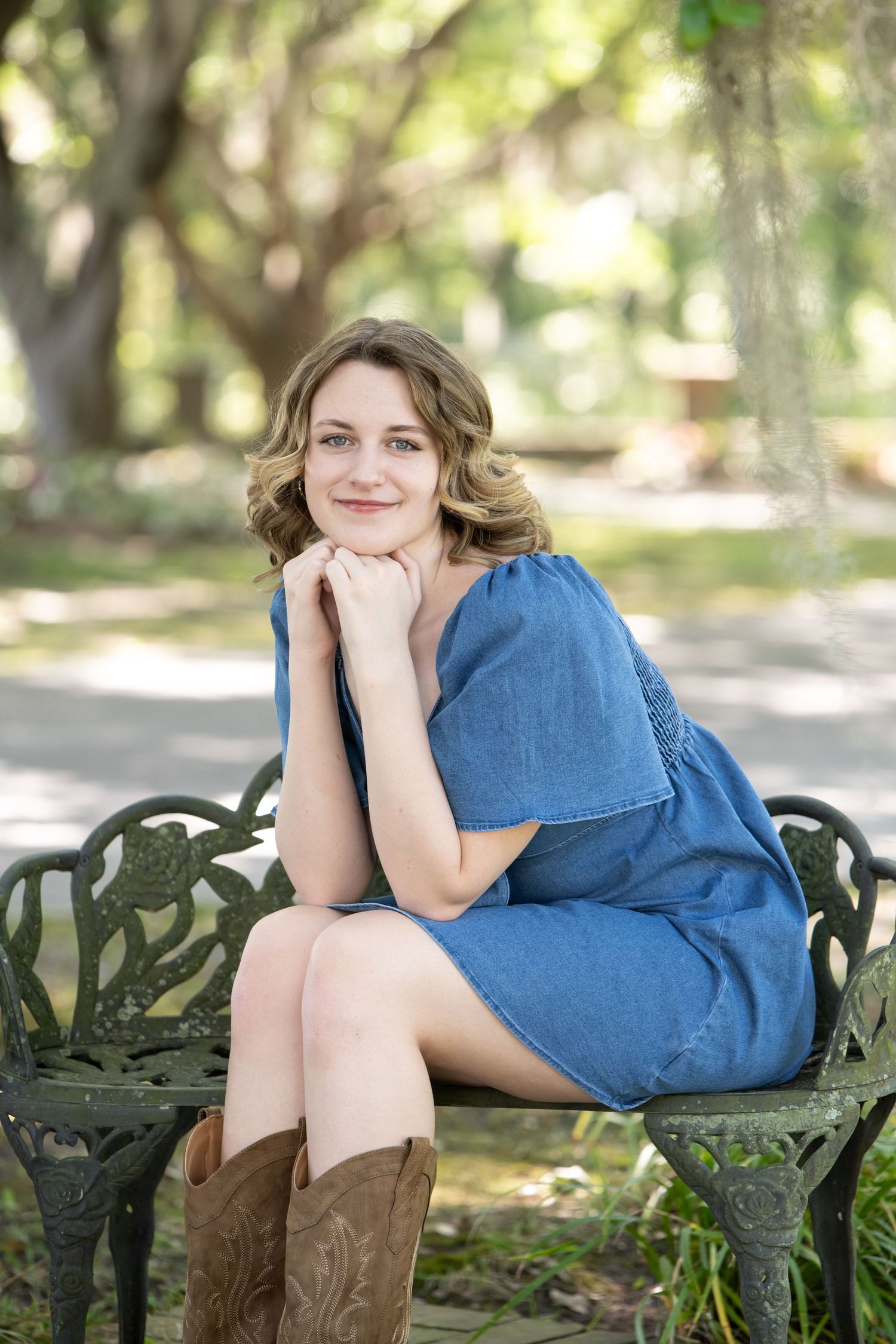 Young woman in denim dress and cowboy boots sitting on ornate iron bench in lush green park, smiling warmly.