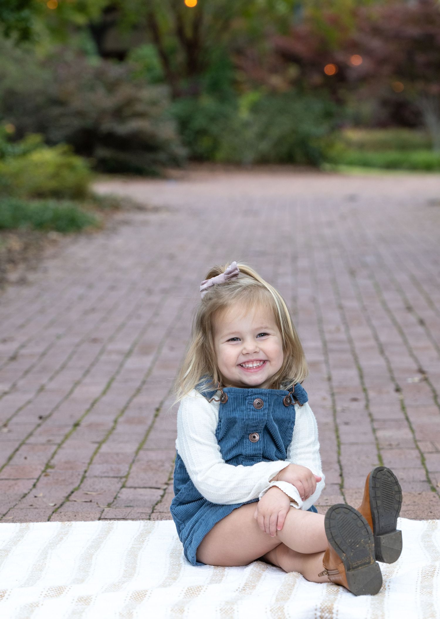 Smiling toddler girl in denim overalls and white shirt sitting on a blanket on a brick path in a garden.