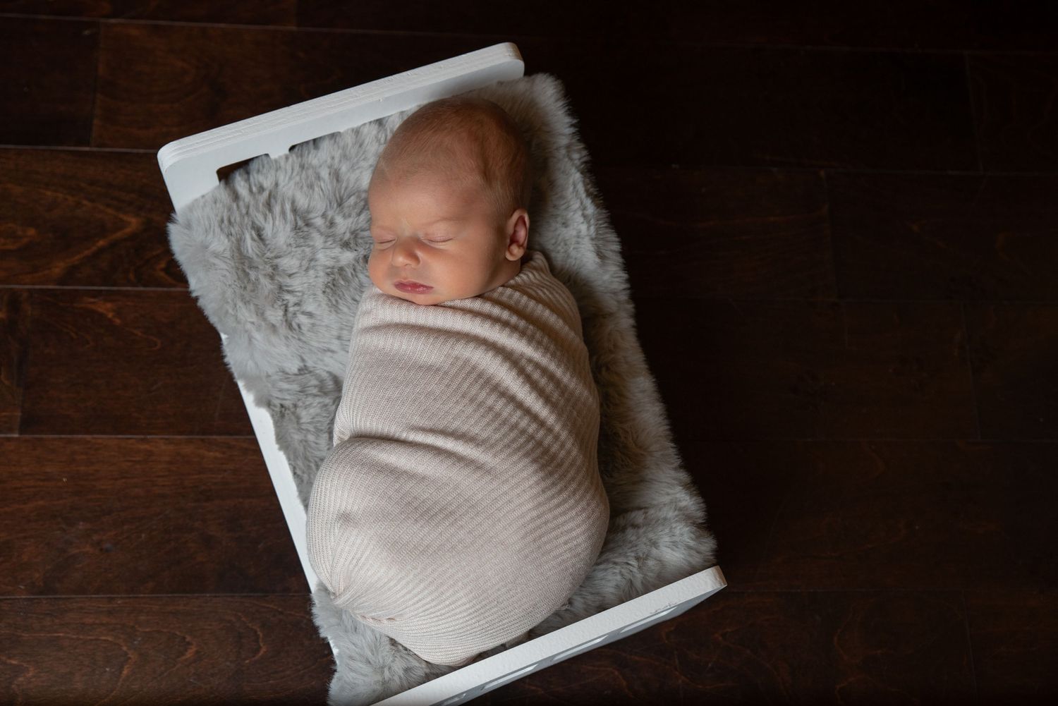 Newborn baby swaddled in cream wrap sleeping peacefully in white wooden box with gray fur lining.