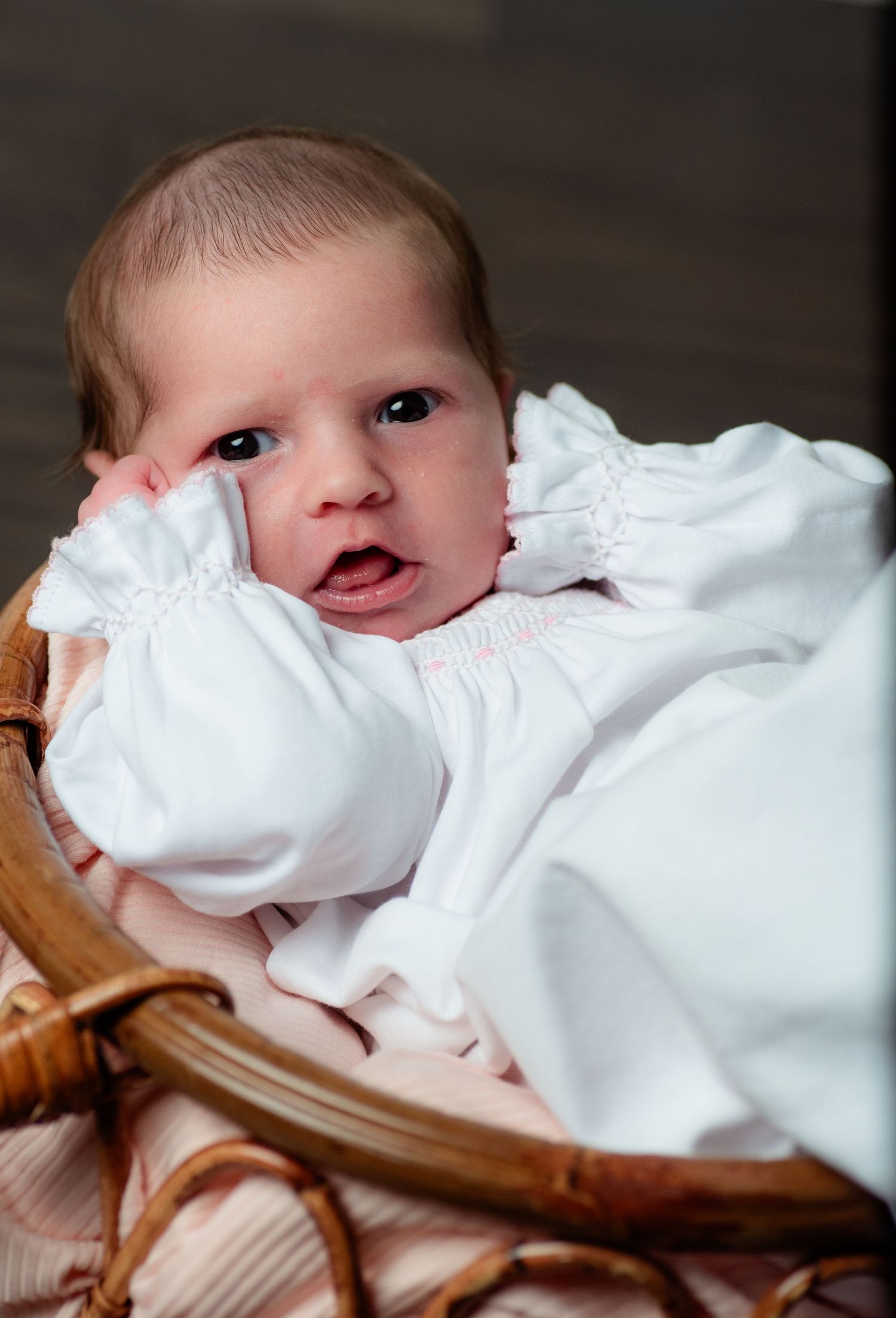 Newborn baby in white outfit resting in a wicker basket, looking alert with hand near face.