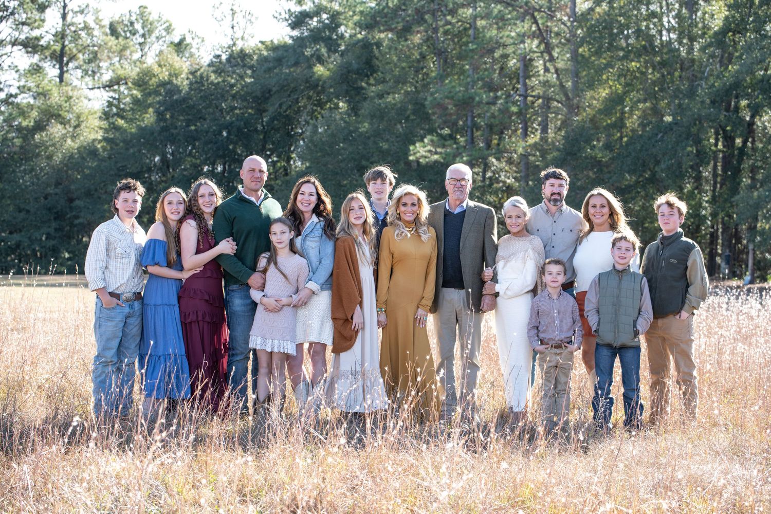 Large family group portrait outdoors in golden field with tall pine trees, multi-generational gathering in coordinated outfits.