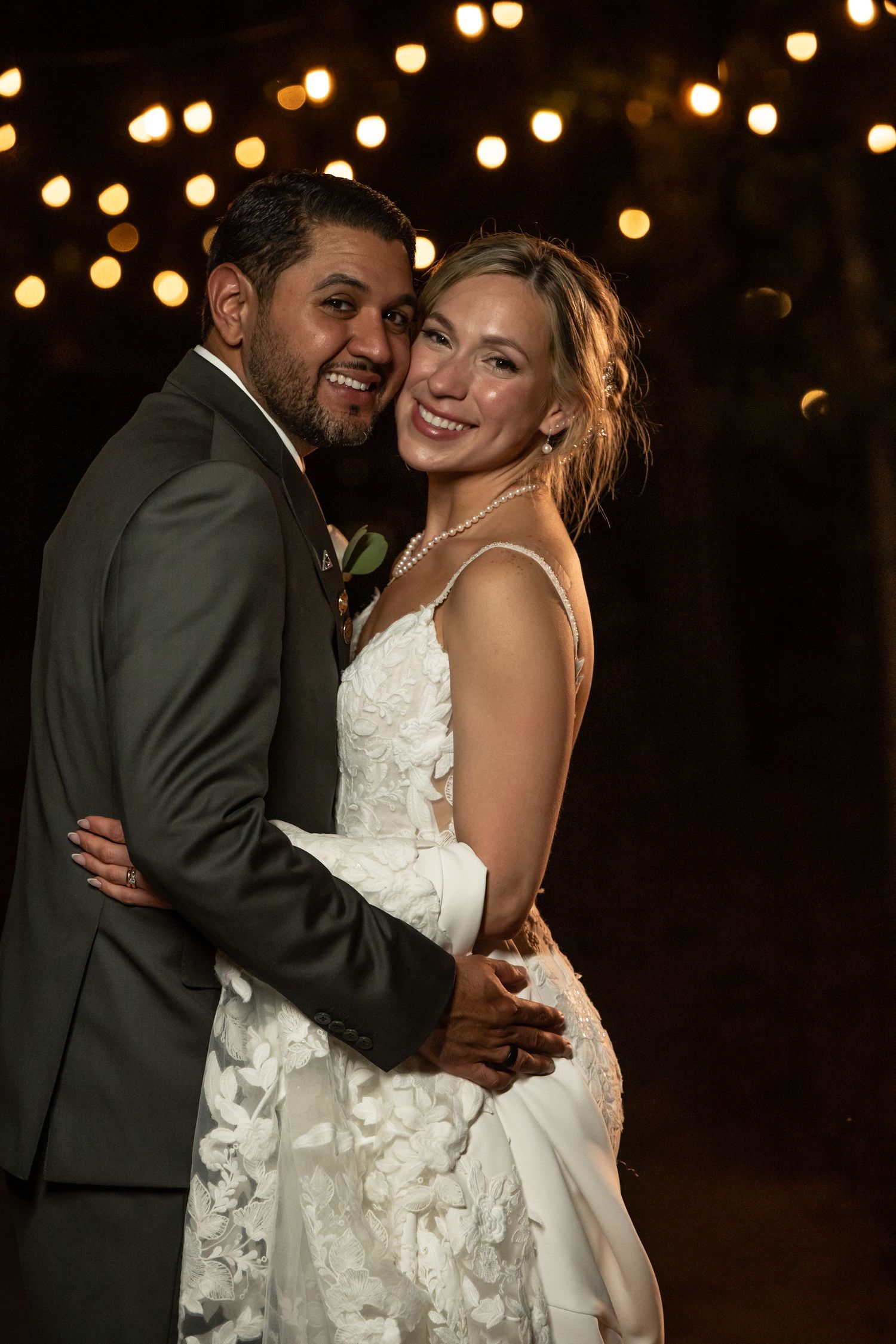 A smiling bride and groom embrace outdoors at night under warm string lights at their wedding reception.