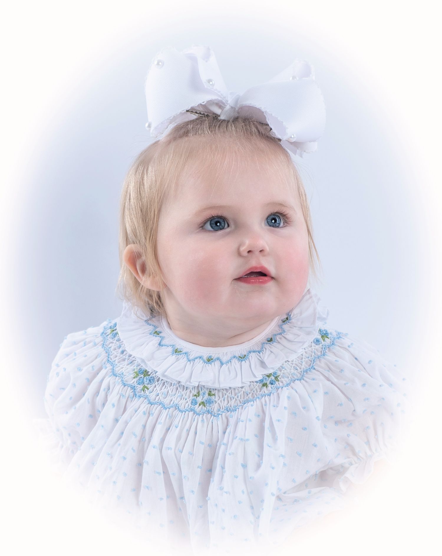 Baby wearing delicate white smocked dress with blue embroidery and white bow against soft vignette background.