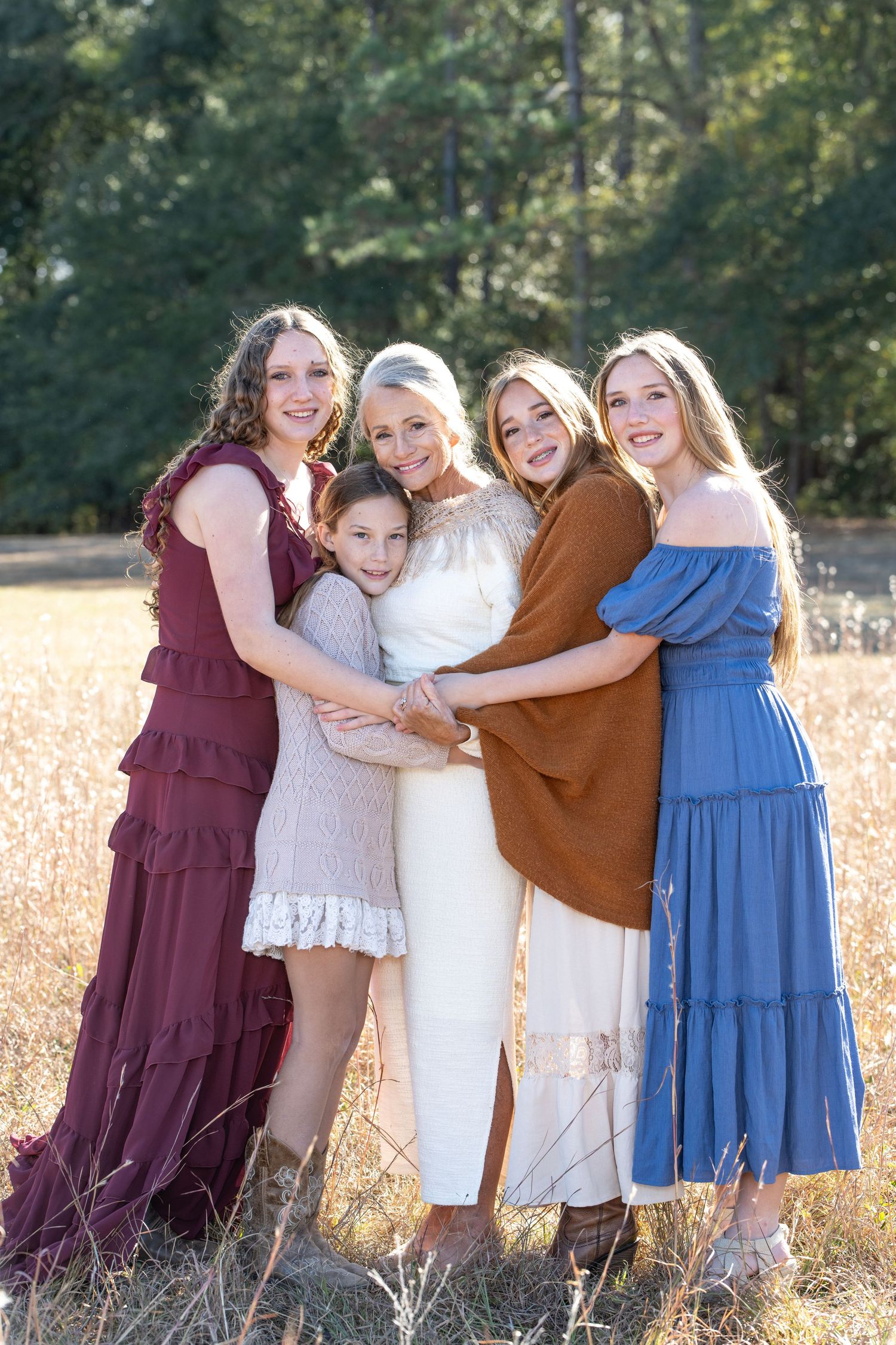 Five women and girls in colorful dresses pose together in a sunlit field, smiling warmly in a family portrait.
