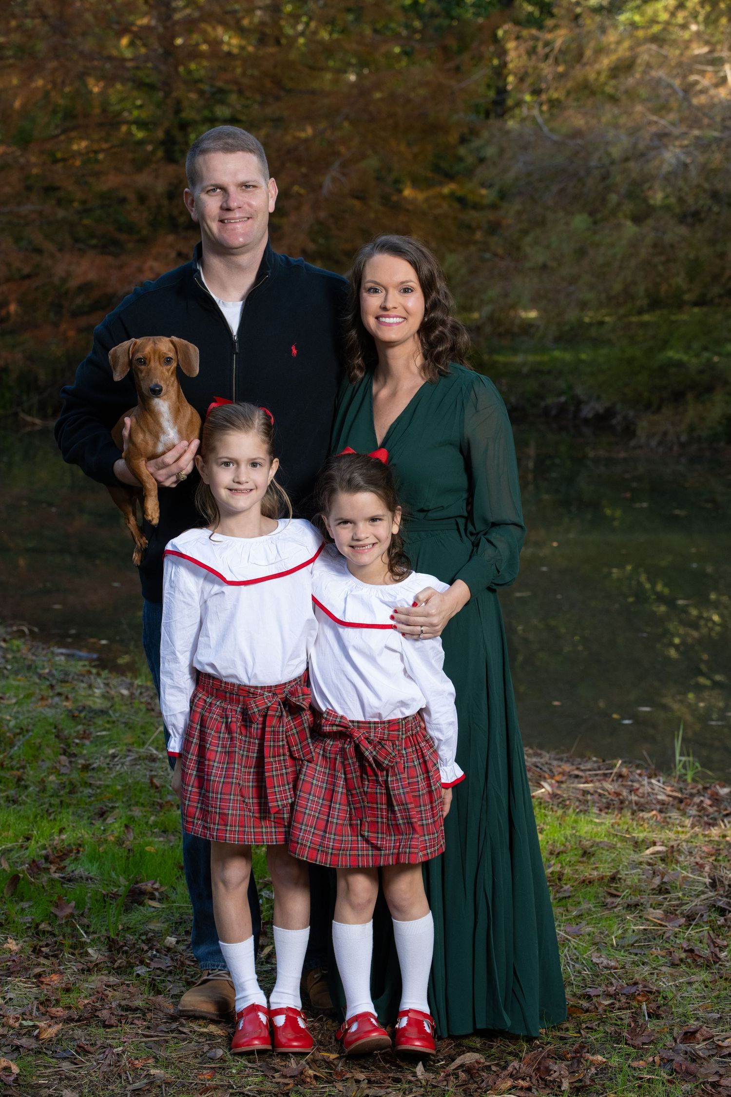 Family portrait with two girls in plaid skirts, parents, and a dachshund dog posed outdoors near a pond in fall.
