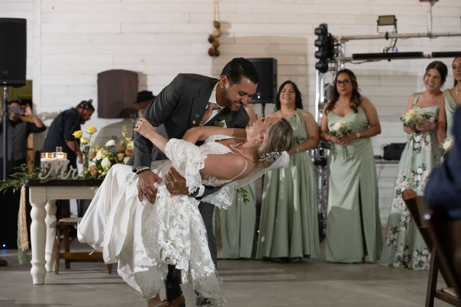 Groom dips bride during first dance at rustic wedding reception with bridesmaids in sage green dresses in background.