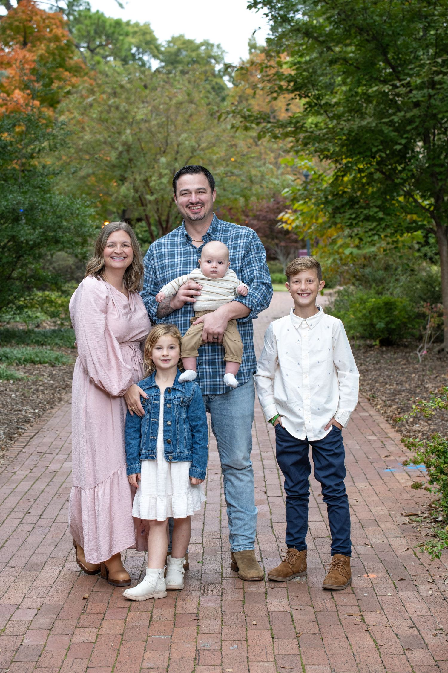 Family of five posing on brick path surrounded by autumn trees, parents with three children including a baby.